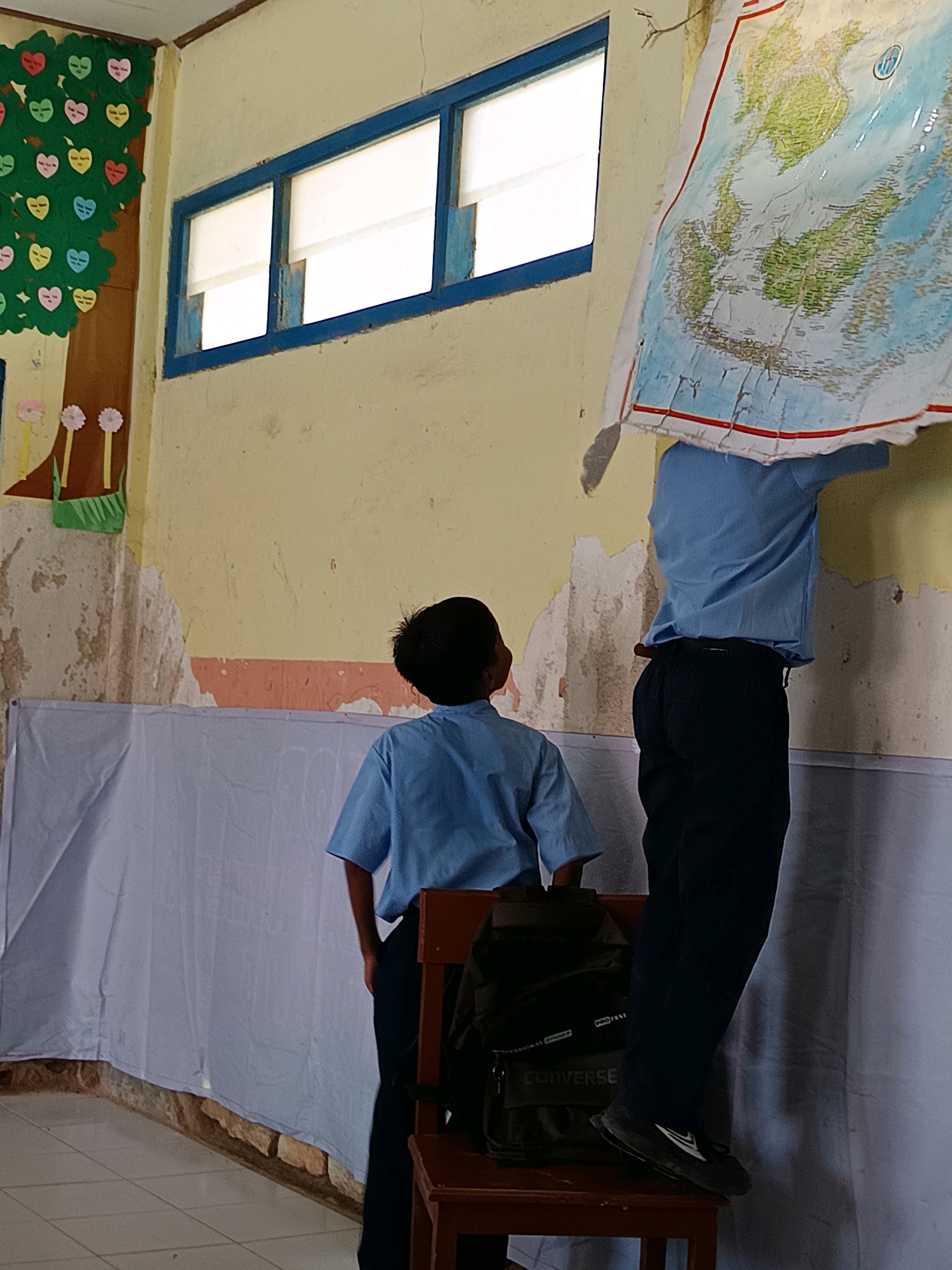 In a classroom in Labuhan, Indonesia, two young boys in light blue uniforms are seen. One boy stands with his back to the camera, facing a wall decorated with a colorful tree of hearts. The other boy is positioned on a chair, holding up a large, creased world map, obscuring his upper body and face. The classroom appears aged, with peeling yellow paint on the walls and a blue-trimmed window with white blinds. A white sheet is draped along the lower portion of the wall, and a backpack rests on the chair the boy is standing on.