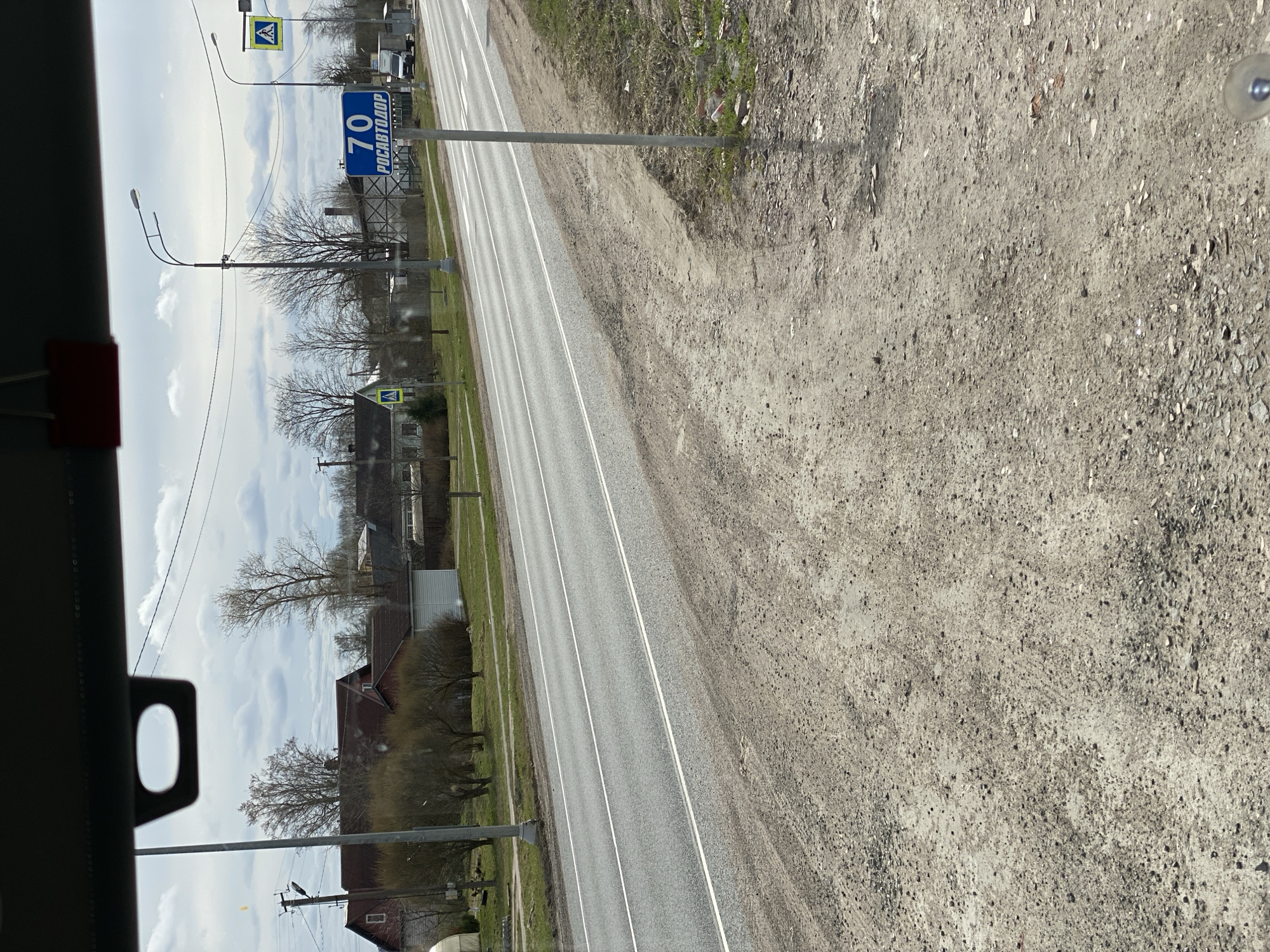 The image captures a daytime scene from inside a moving vehicle, looking forward along a two-lane asphalt road in Bol'shoe Teshkovo, Russia. The road features clear double white lines down its center.
To the left of the road, a green grassy verge separates the roadway from a semi-rural landscape. This area contains several bare trees, suggesting late winter or early spring. Various residential buildings with red and grey roofs are visible beyond the trees. Multiple utility poles with streetlights and overhead power lines line this side of the road. Prominent road signs include two yellow diamond-shaped signs, each depicting a black pedestrian crossing symbol. A blue rectangular sign clearly displays "70" in large white digits, along with the text "РОСАВТОДОР" (ROSAVTODOR), which is the Russian Road Agency, likely indicating a speed limit of 70 km/h or a road designation.
To the right of the road, a wide, unpaved shoulder of dirt and gravel stretches into the foreground, showing textured ground with small stones.
The sky is partly cloudy with bright light, casting faint shadows. A dark interior element with an oval cutout is visible in the bottom left of the frame, confirming the perspective from within a vehicle. The scene depicts a typical, calm road environment.