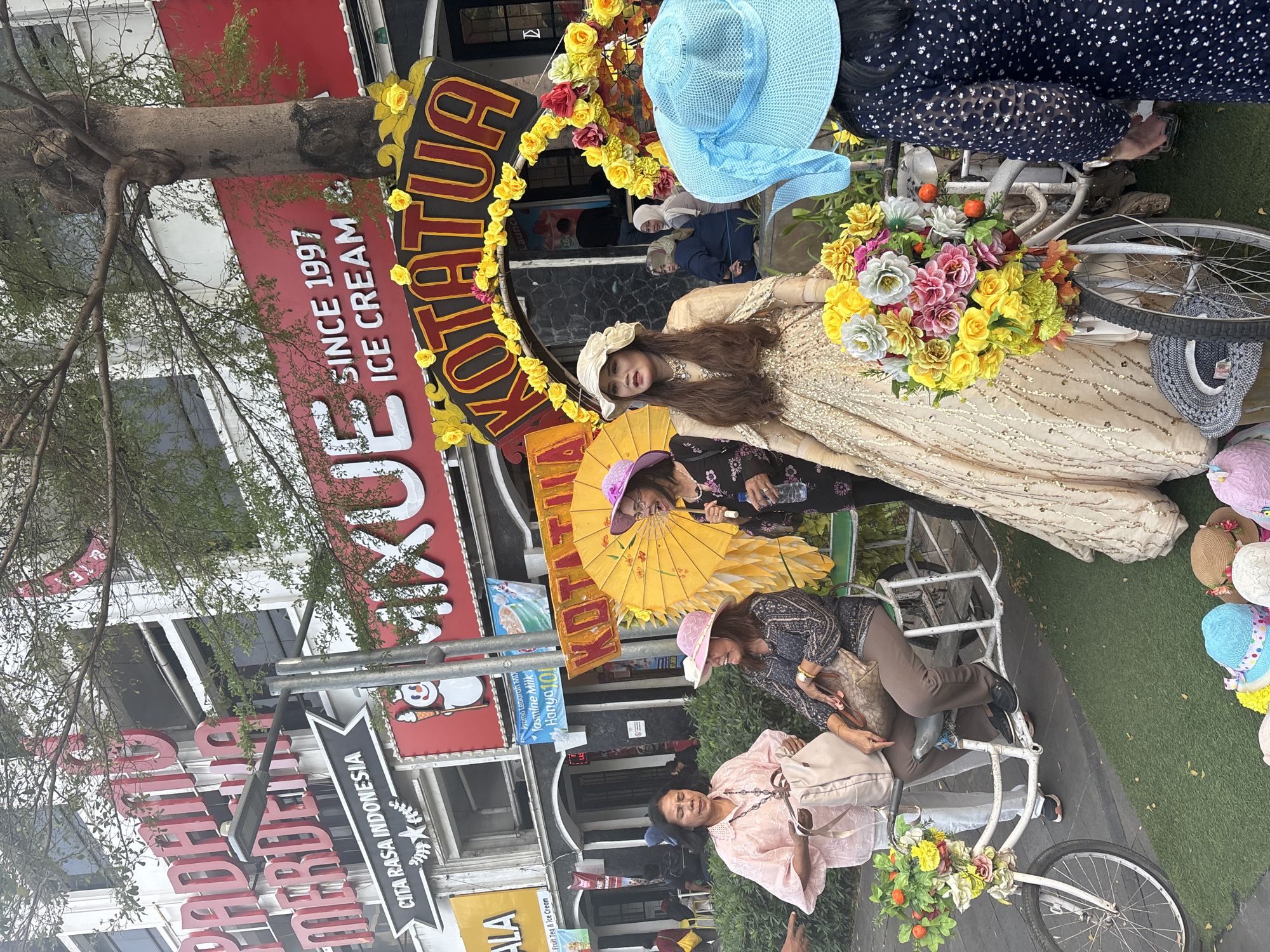 This image captures a street scene in Kota Tua, West Jakarta, Indonesia, likely near the Fatahillah Square area, indicated by the decorative arch and the general atmosphere. A group of people, primarily women, are gathered around a white bicycle decorated with a large bouquet of colorful artificial flowers. One woman, dressed in a shimmering gold gown and a wide-brimmed cream hat, stands prominently in the foreground, posing for the camera.
To her left, two women are seated on a similar decorated bicycle. The woman in the pink blouse holds a large beige bag, while the woman beside her wears a dark patterned top and a pink hat. Another woman, in a black dress and a purple hat, stands to the right of the seated women, holding a yellow umbrella and a bottle. In the background, several shops are visible, including one with a large red sign that reads "MIXUE ICE CREAM" and "SINCE 1997". Another sign in red letters says "PADANG MERDEKA". The architecture suggests a historic urban setting. The ground is covered with artificial grass in some areas, and several hats are displayed on the ground in the foreground, suggesting a place for tourists to take photos or purchase souvenirs. The overall scene is vibrant and suggests a tourist attraction or a festive event.