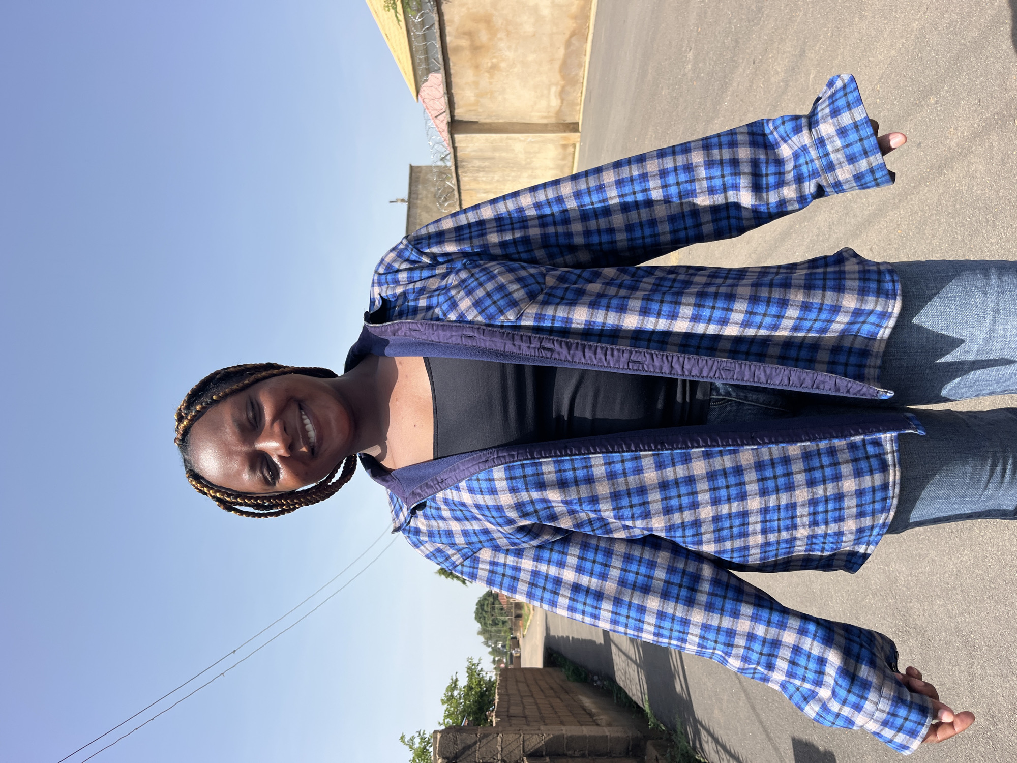 A medium shot captures a young Black woman standing outdoors on an asphalt road, with buildings and walls visible in the background. She is facing forward, with her body slightly angled and her arms extended outwards. The woman has her eyes closed and is smiling broadly, with her teeth showing. Her dark hair is styled in braids with some lighter streaks. She is wearing a black tank top underneath an open blue, grey, and white plaid long-sleeved shirt. Her lower body is clad in blue jeans. The environment appears to be an urban or suburban setting under a clear blue sky, with a brick wall and a street on the left, and a tan stucco wall with barbed wire on the right. The sun is casting shadows, indicating it is daytime.