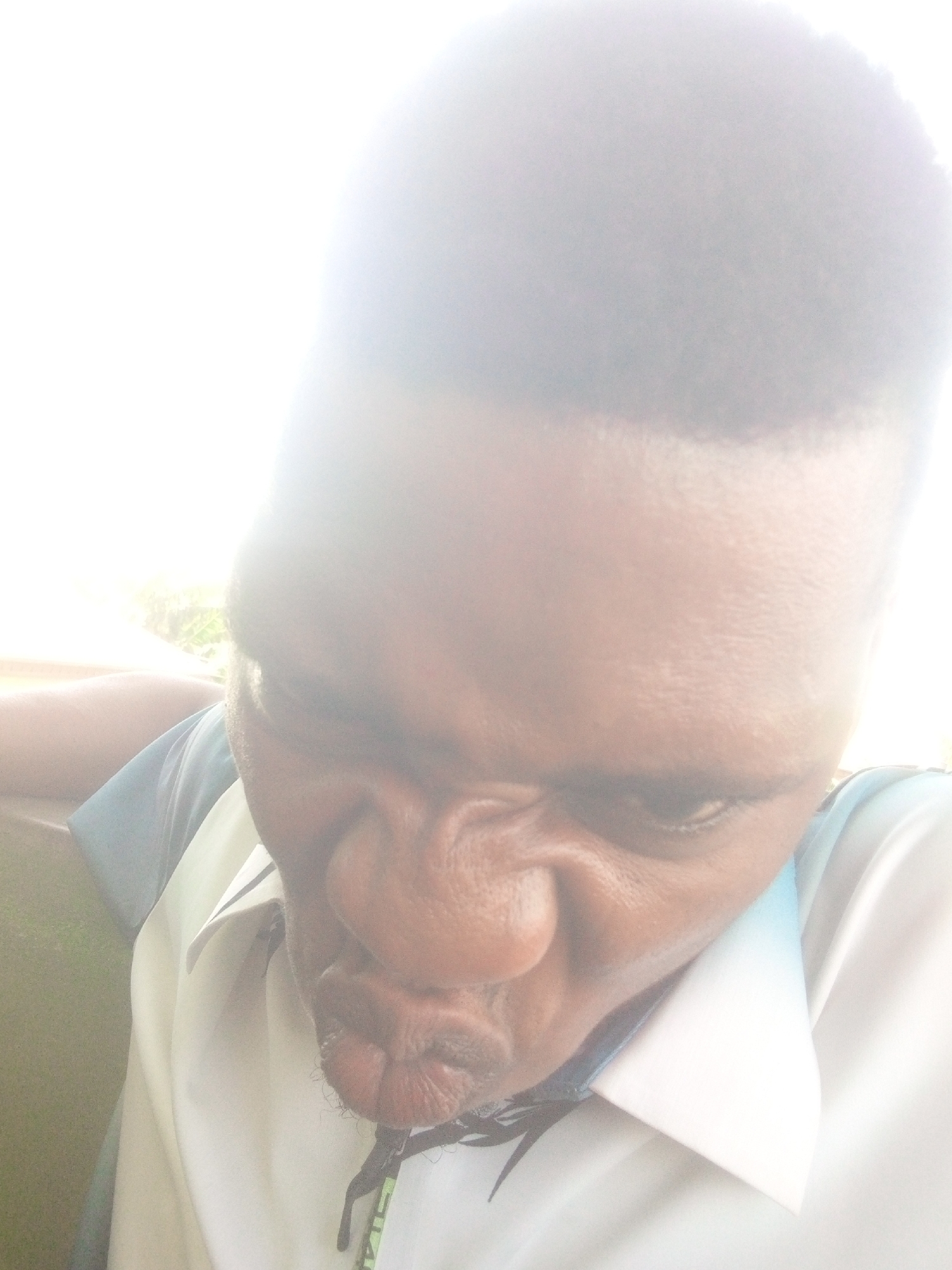 A person with dark skin and short, dark hair is looking down at the camera with pursed lips. The person is wearing a white shirt with light blue accents on the shoulders and collar. The lighting is very bright, obscuring the details of the face and background. The location is unknown.