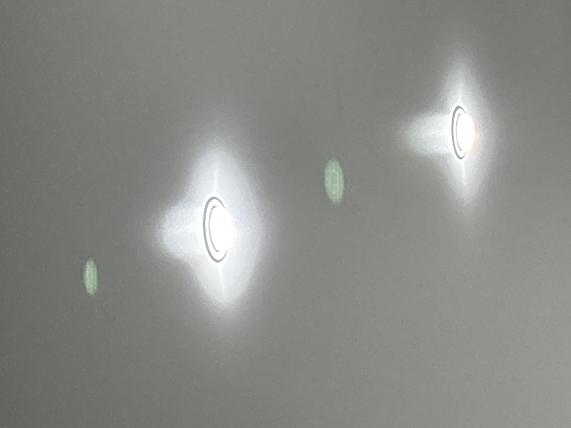 The image shows a light gray surface with four distinct light reflections. Two of the reflections are bright, circular shapes with dark semicircular lines, and radiating light. The other two are fainter, elongated, and greenish. The setting and location are unknown.