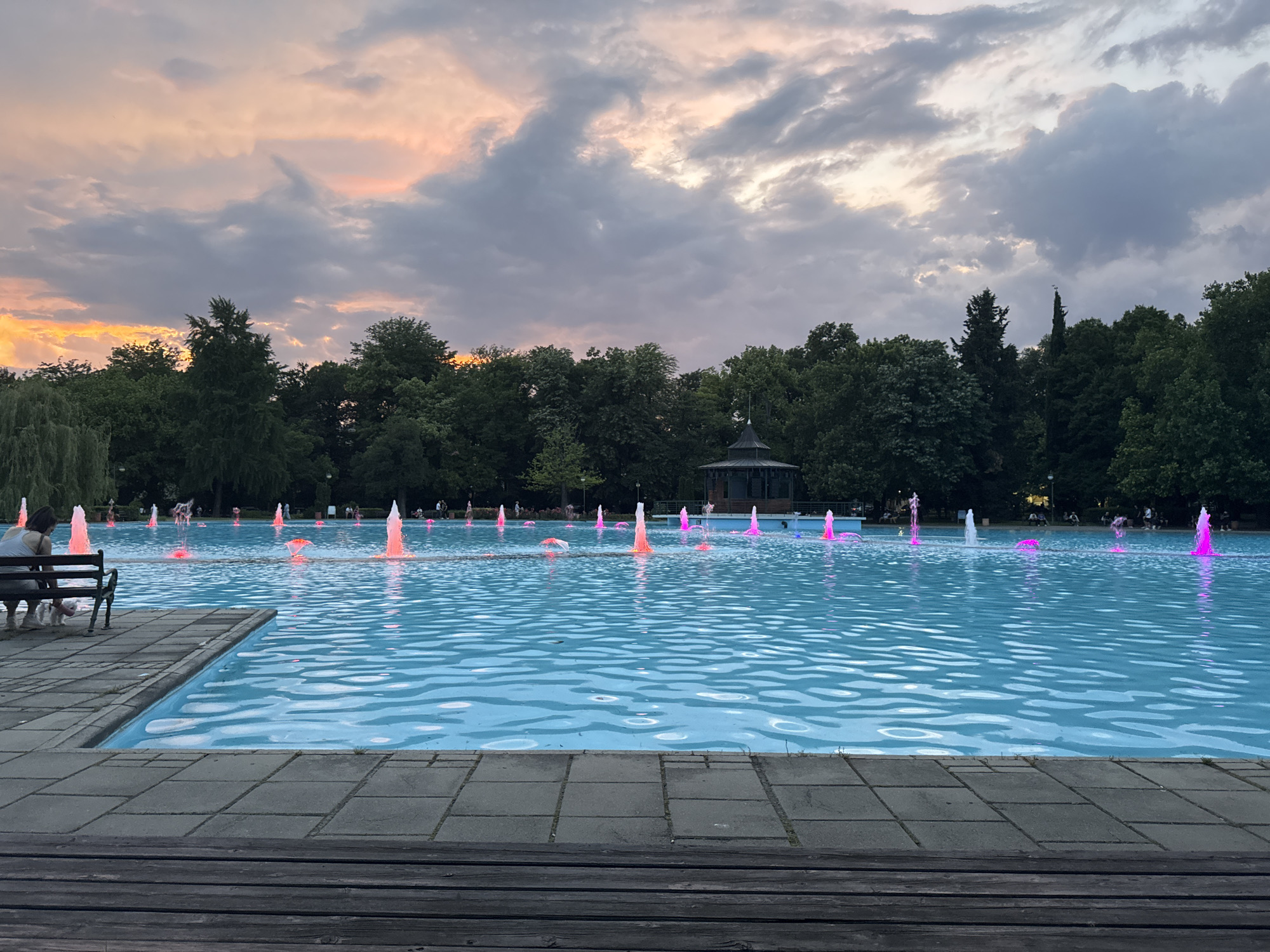 The image depicts the Singing Fountains in Plovdiv, Bulgaria, during twilight. A large, rectangular pool of water, illuminated by colorful underwater lights, dominates the foreground and midground. The water surface reflects the sky and the illuminated fountains. Numerous fountains are active, emitting streams of water that are lit in vibrant pink, orange, and white hues. In the left foreground, two individuals are seated on a park bench beside the pool. The background features a line of lush green trees, beyond which a dark, pavilion-like structure with a pointed roof is visible. The sky above is filled with dramatic clouds, tinged with the warm colors of the setting sun, ranging from pale orange to shades of grey.