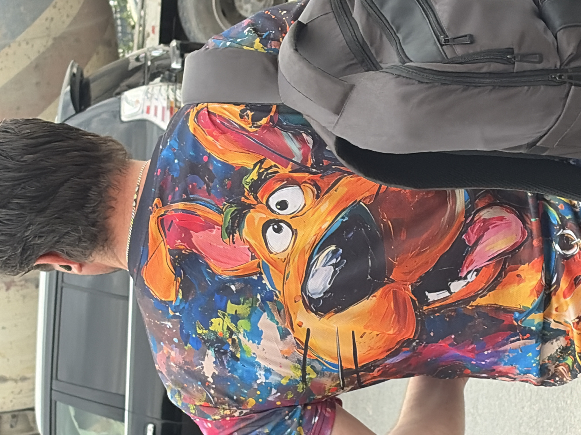 A person seen from behind is wearing a colorful t-shirt with a graphic of Scooby-Doo. They are also wearing a gray backpack and have earbuds in their ears. The person is standing outdoors, with a black car and a construction vehicle visible in the background.