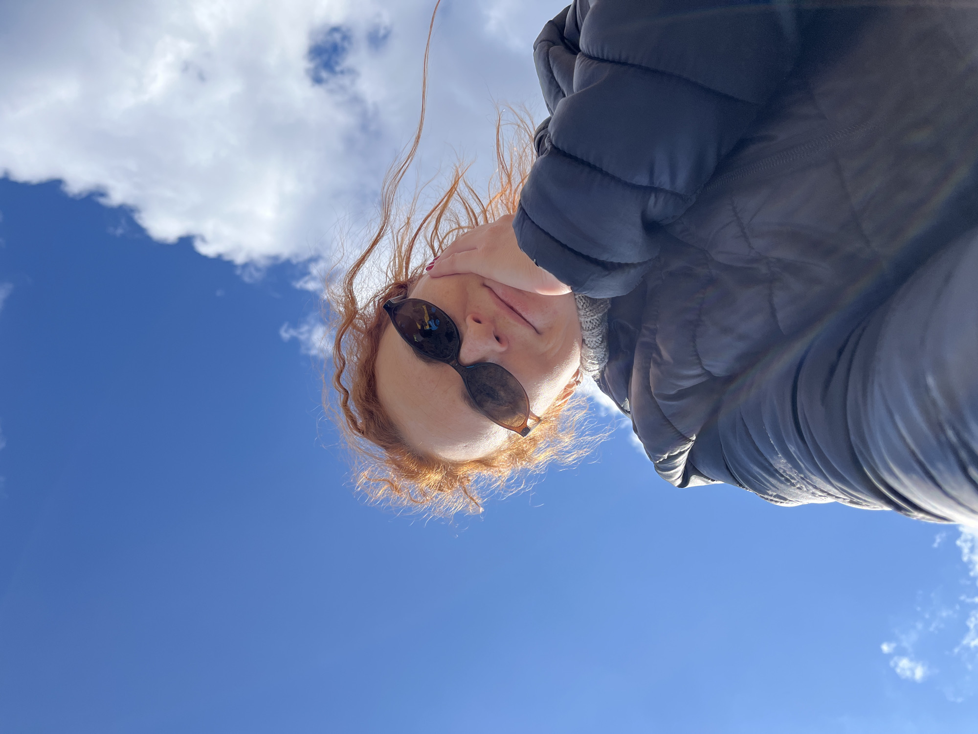 A low-angle, close-up shot depicts a person wearing sunglasses and a dark-colored puffer jacket, with a hand near their face. The background consists of a blue sky with some clouds. The location, Chaikovsky, Russia, is not discernible from the image itself.