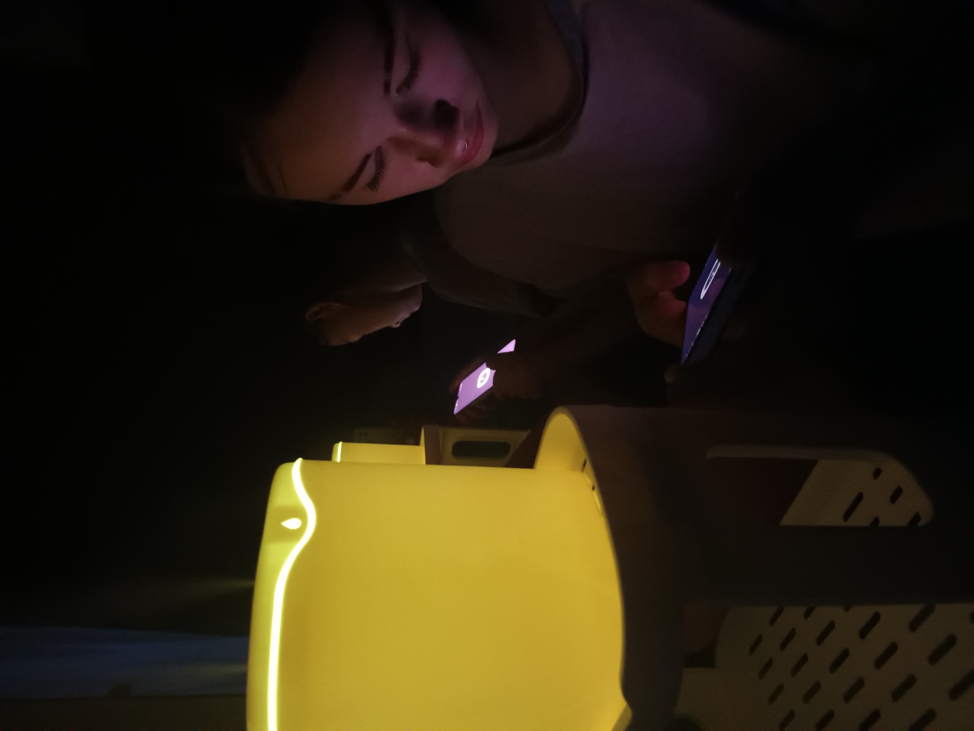 A woman is reclining in a public space in Ольховка, Russia. She is looking at her phone. Several illuminated structures are also visible, seemingly part of a public installation or seating arrangement.