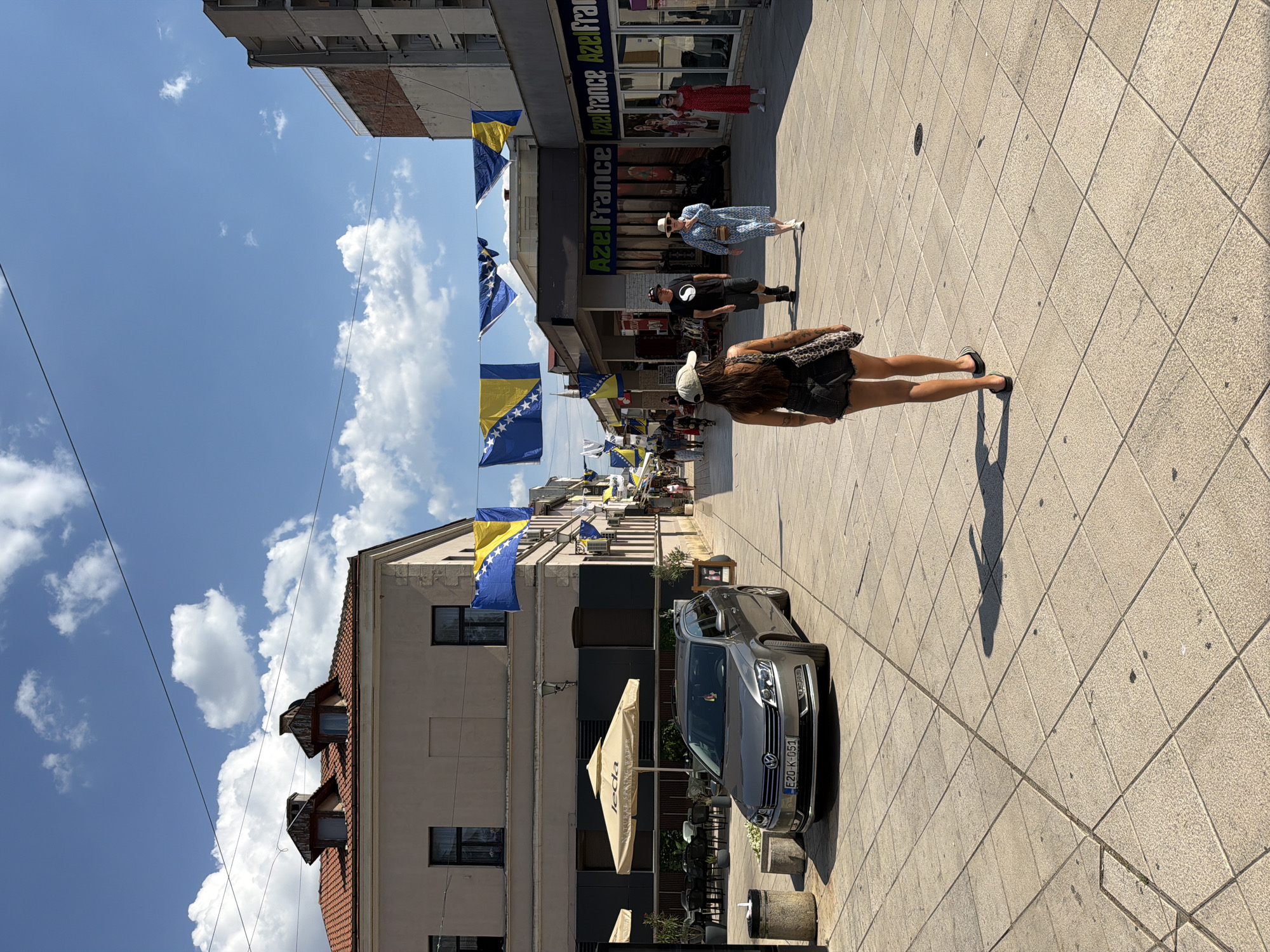 The image depicts a street scene in Mostar, Bosnia and Herzegovina, with Bosnian flags strung across the street, indicating a national celebration or event. The street is paved with light-colored square tiles, and on the left, a silver Volkswagen Passat wagon is parked in front of a building with awnings and outdoor seating. The building on the left has a traditional facade with a tiled roof and two dormer windows. On the right side of the street are several buildings with storefronts, one of which has the name "AzelFrance" visible. Several people are walking along the street. In the foreground, a woman wearing a black tank top, denim shorts, and a baseball cap is walking away from the camera. Behind her, a man in a black t-shirt and shorts walks with a woman in a blue floral dress. Further down the street, more people are visible, including a woman in a red dress. The sky is blue with scattered white clouds.