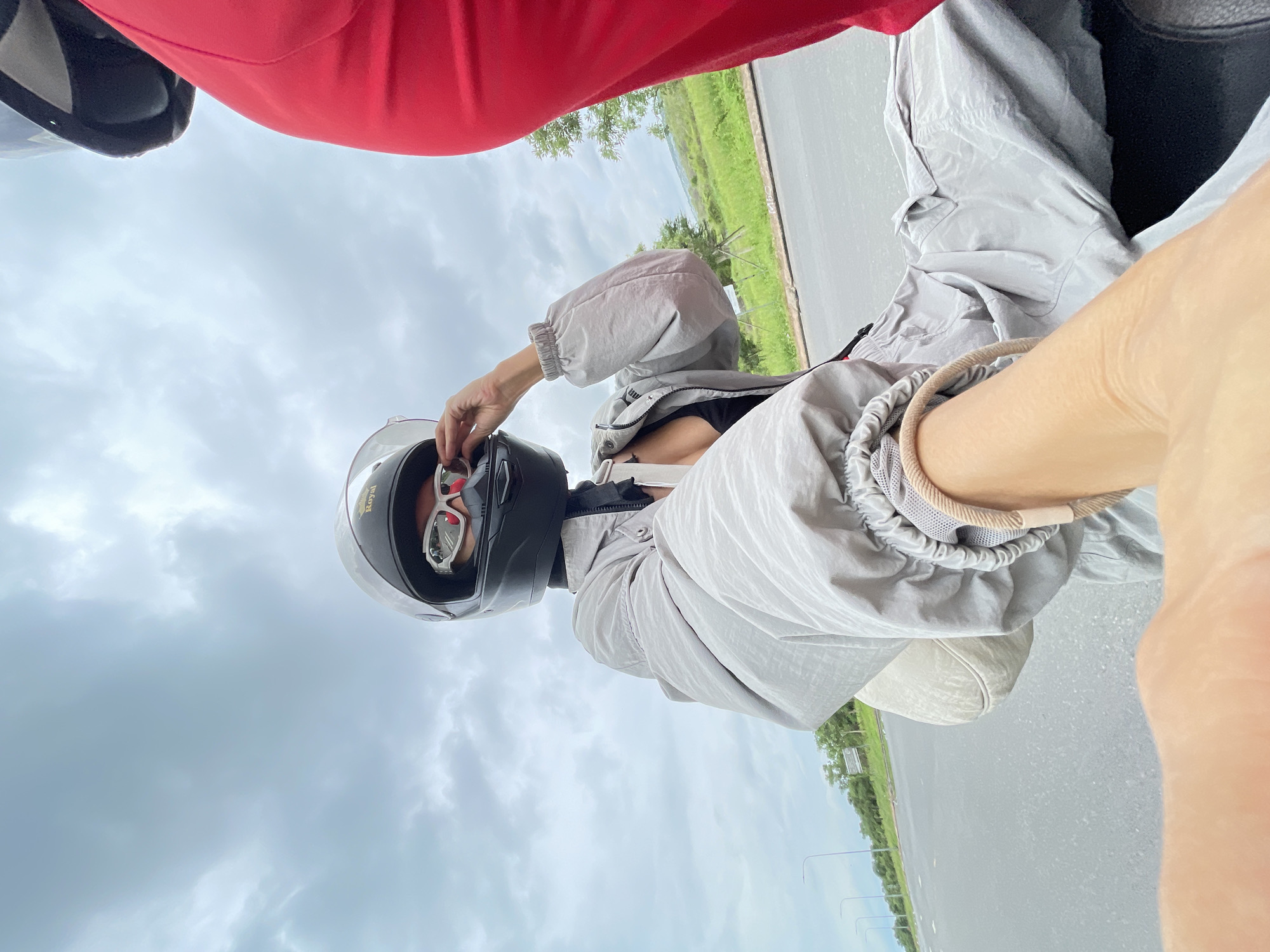 The image is a selfie taken from the perspective of a person riding on the back of a motorcycle. The rider in the foreground is wearing a black full-face helmet with white goggles, a grey windbreaker jacket, and a black top. They are adjusting their goggles with their right hand. The motorcycle is moving on a paved road, likely in a rural or suburban area, with grassy shoulders and trees visible in the background. Another person, partially visible on the right side of the frame, is wearing a bright red garment, possibly pants or a skirt. The sky is overcast. The location is identified as Phu Thuy Ward, Vietnam.