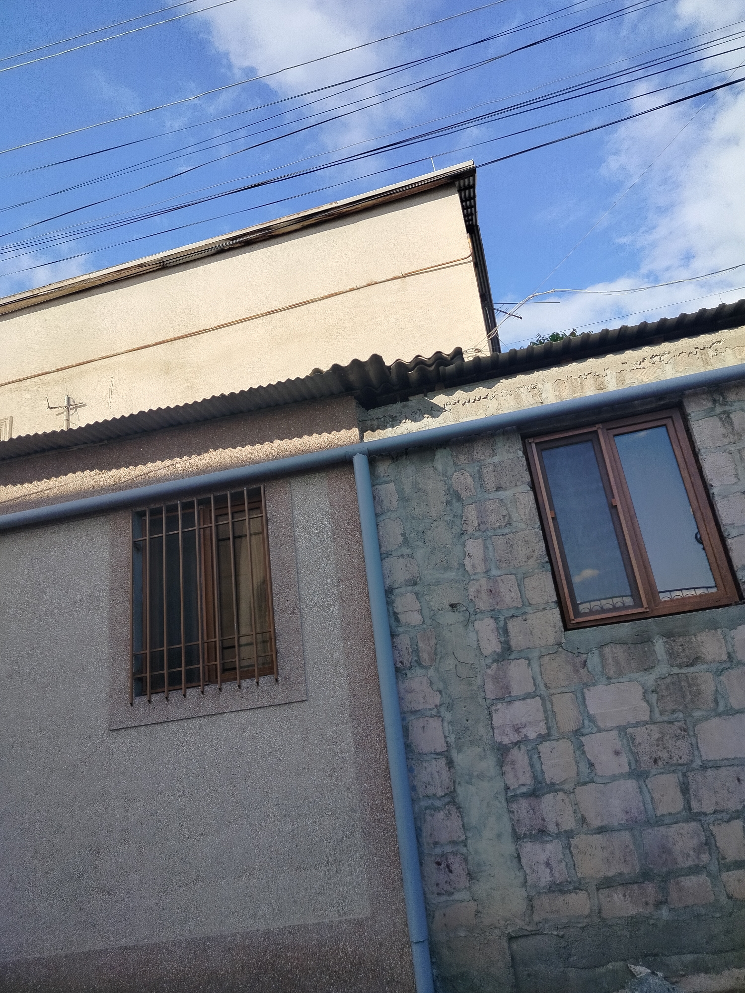 The image shows two adjacent buildings in Yerevan, Armenia. The building on the left is plastered in a light-colored stucco and has a window with metal bars. The building on the right is made of exposed brick and has a double-paned window. A gray pipe runs vertically between the two buildings. Power lines are visible against a blue sky with clouds in the background.