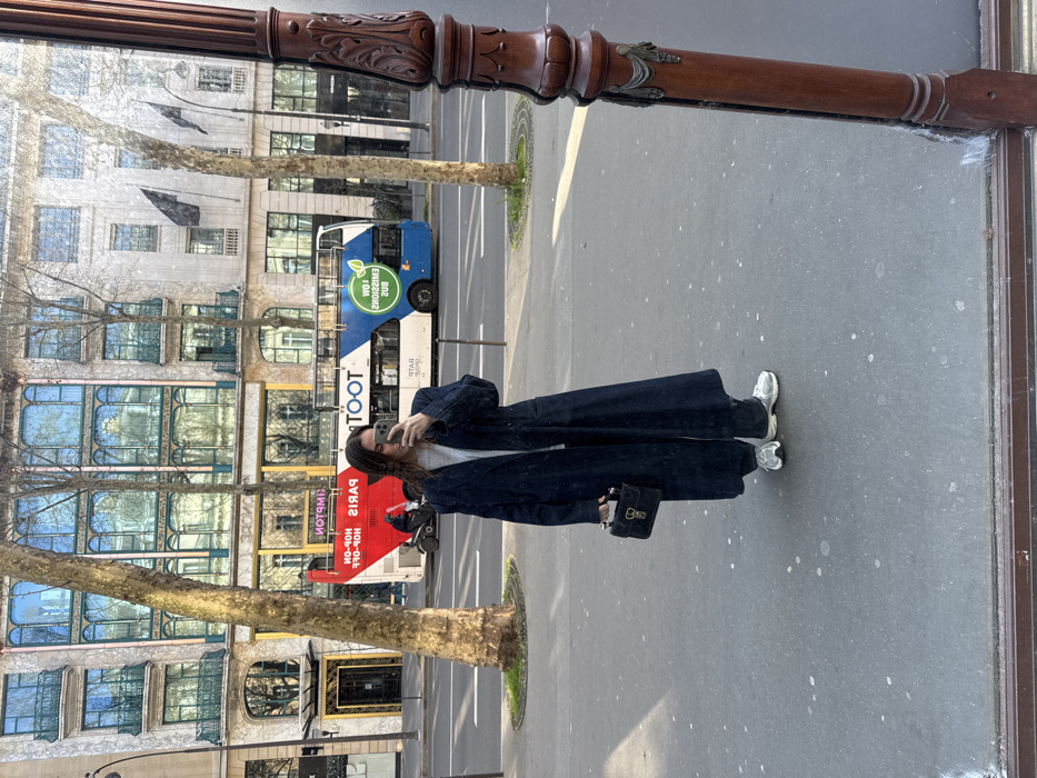 The image captures a female subject reflected in a mirror or large window on a Parisian street. The subject, positioned centrally, has long dark hair and is dressed in a light-colored top, a long dark overcoat, dark trousers, and light athletic shoes. She holds a small dark handbag with a metallic clasp in her left hand and a smartphone in her right, actively taking the photograph.
Behind the reflection, a red and blue double-decker "Hop-On Hop-Off PARIS" tour bus is visible on the street, with its lettering mirrored. A scooter rider is discernible near the bus. Across the street, classic Parisian buildings with ornate facades, large windows, and balconies line the boulevard. One building on the left features distinct arched windows and green railings. Bare deciduous trees are spaced along the sidewalk. The scene is illuminated by natural daylight. A decorative wooden post, part of the mirror's frame, is visible on the right edge of the image. The specific architectural style and presence of the tour bus strongly suggest a location on a prominent boulevard in Paris, France, possibly near a major department store like Printemps or Galeries Lafayette on Boulevard Haussmann.