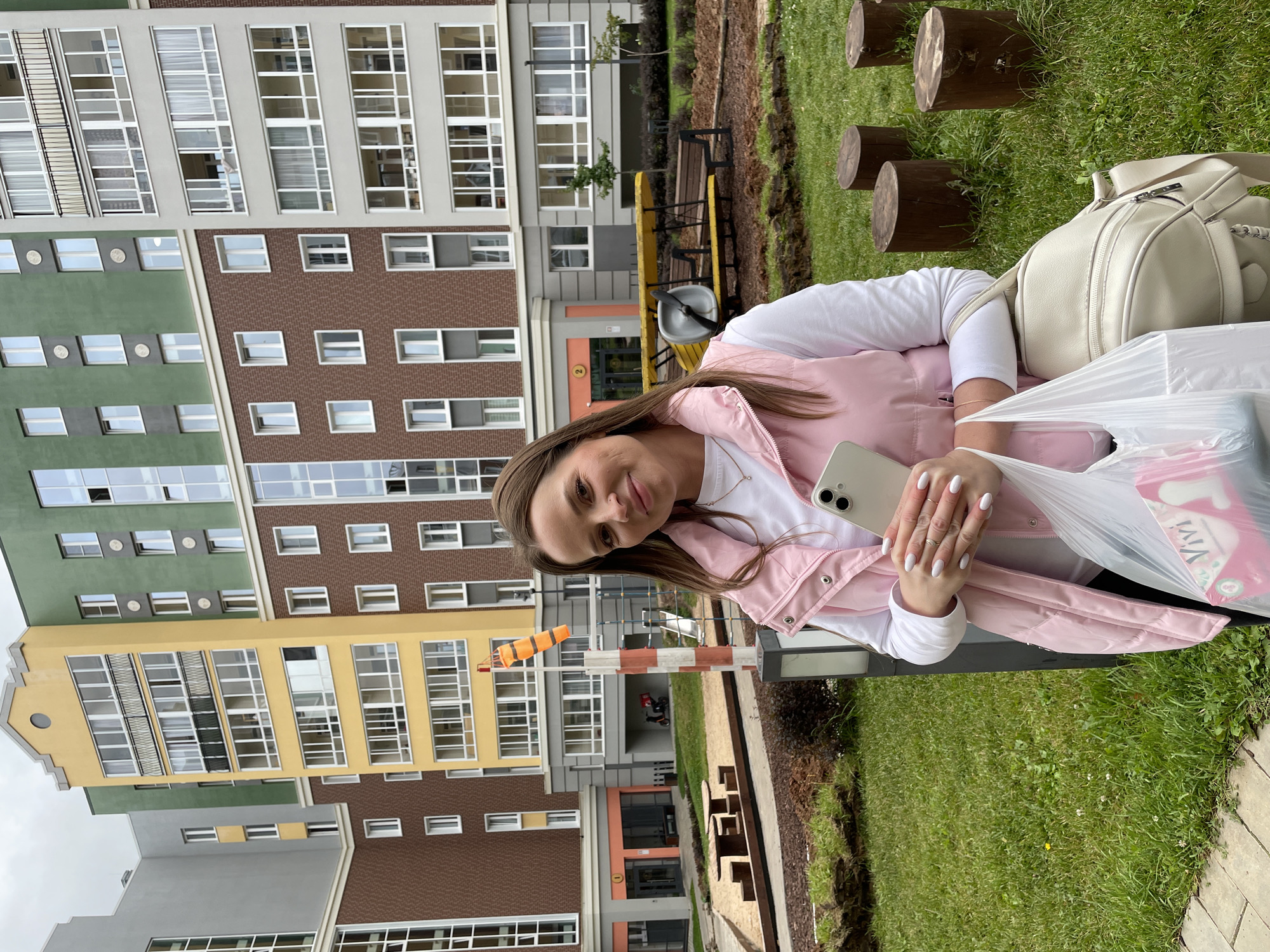 In Pervomaysky, Russia, a woman with long brown hair is standing on a grassy area in front of a modern apartment complex. She is wearing a white long-sleeved shirt and a pink vest. In her right hand, she is holding a smartphone, and in her left, she is carrying a white plastic bag. Beside her, on her right, is a light-colored backpack. The apartment building features a multi-colored facade with yellow, green, and brick sections, and numerous windows. In the background, there is a playground area with wooden benches and a small airplane-shaped structure. The ground is covered in grass, with several short, thick tree stumps scattered around. A weather vane with orange and white stripes is visible on a pole in the middle ground.
