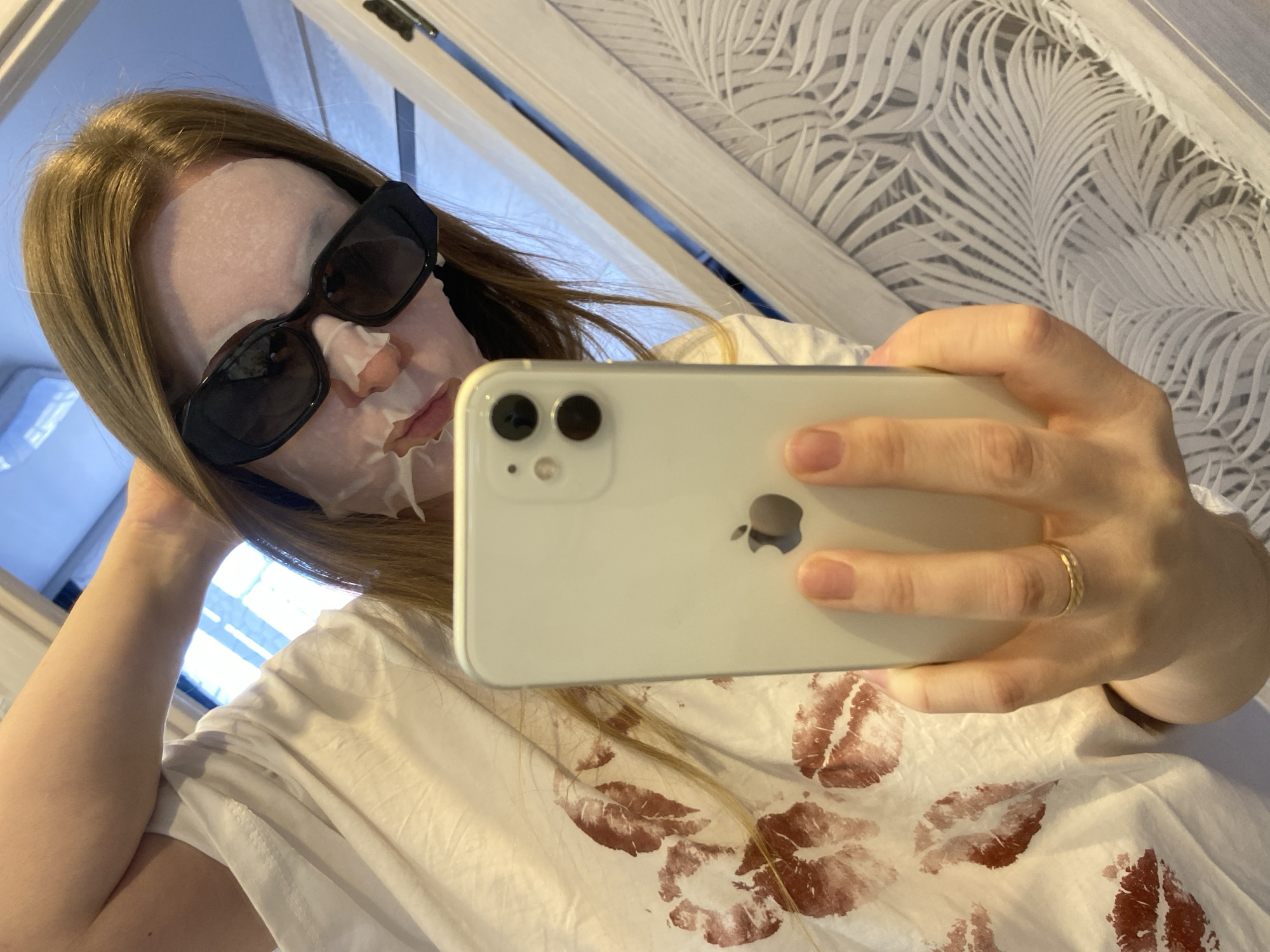 A person is taking a selfie indoors, wearing a white face mask and oversized black sunglasses. The person is holding a white smartphone in their right hand, displaying the phone's back which includes the Apple logo. The person is wearing a t-shirt with red lipstick kiss prints. The background features a white wall with a leafy pattern. The image seems to have been taken at an angle with a window or light source visible. No specific details indicate the city of Noviy, Russia.