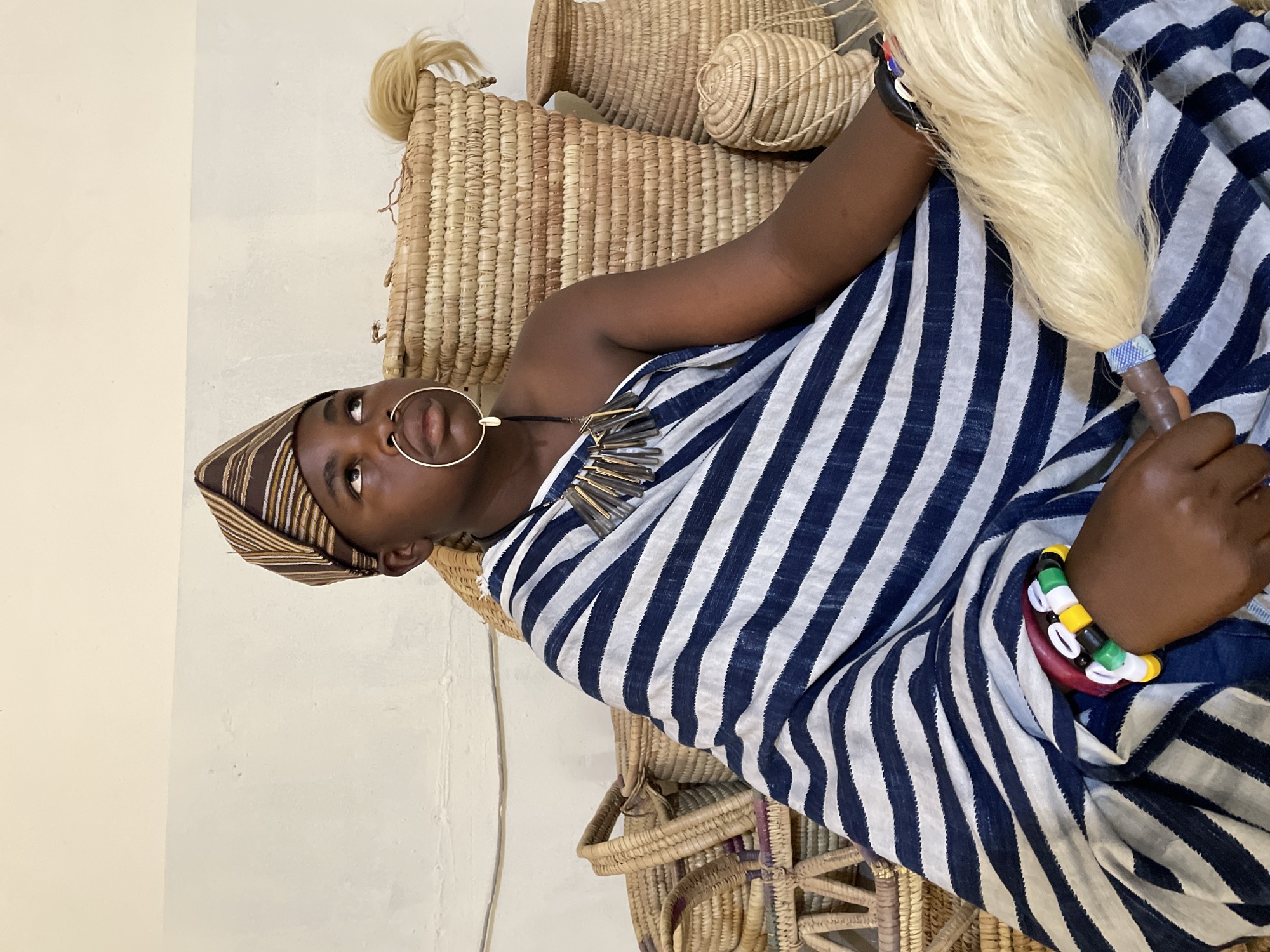 A person is dressed in a striped navy and white cloth and hat, holding a bundle of what appears to be flax. The person is sitting in a seat made of woven straw, with woven straw baskets surrounding the subject. The person wears a nose ring, a necklace, and a beaded bracelet. The location is the city of Jos, Nigeria.