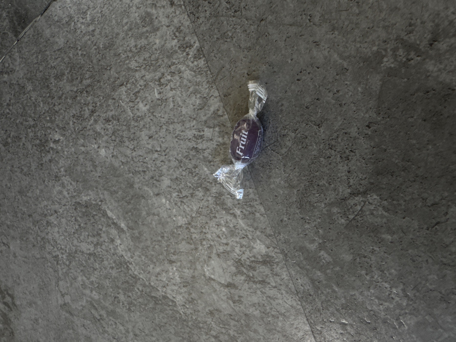 This image depicts a singular, individually wrapped candy resting on a dark grey, textured floor surface in Moscow, Russia. The candy is ovate in shape, purple, and encased in a clear, crinkled plastic wrapper that is twisted at both ends. The word "Fruit" is visible in white sans-serif text on the purple surface of the candy, through the transparent wrapper. The floor, appearing to be concrete or large, textured tiles, exhibits a mottled pattern of varying grey tones. A distinct, thin, dark linear seam or joint traverses the lower mid-section of the surface, running diagonally from the lower-left to the upper-right. No individuals, actions, or interactions are present in the scene.