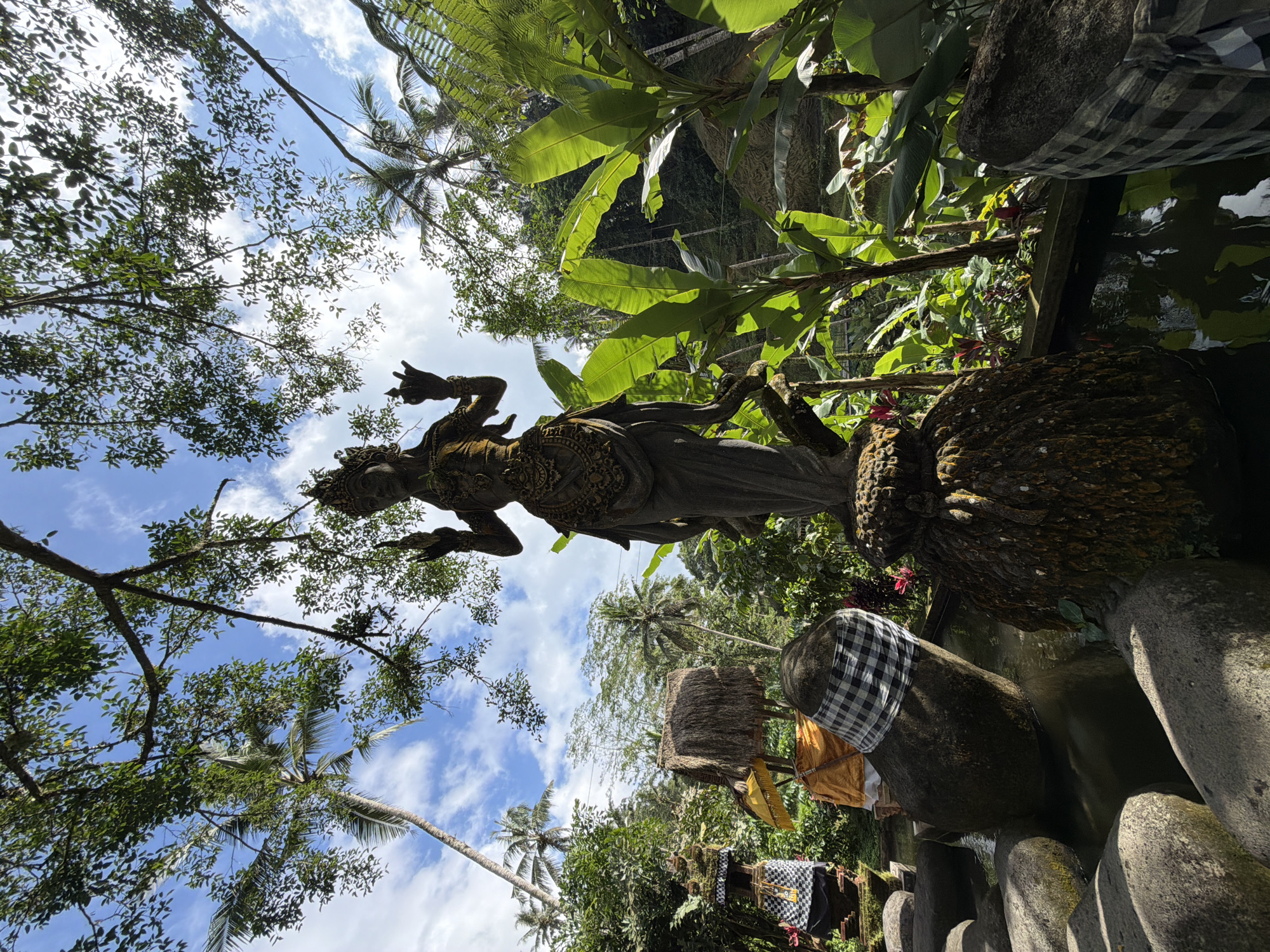 A low-angle shot showcases a statue set amidst lush vegetation and a blue sky. The statue, weathered and ornate, is positioned near a small body of water, with stones decorated with black and white checkered cloths along the bank. The vegetation includes a mix of tropical plants and trees, providing a dense backdrop. The sky is partly cloudy. The location is Tegal Suci, Indonesia.