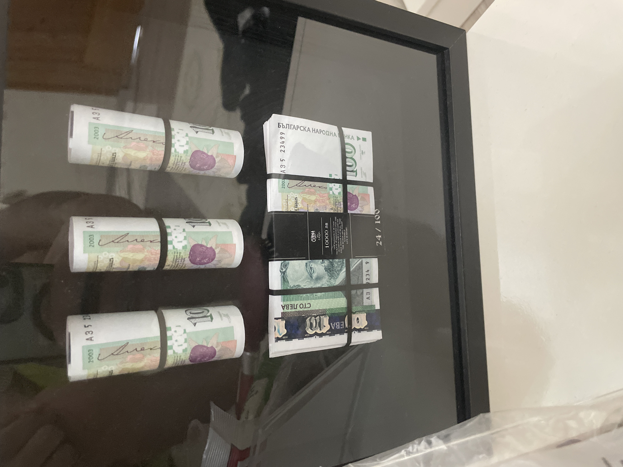 The image displays an arrangement of Bulgarian Lev banknotes, likely presented within a black-framed clear box or display case. Three rolls of 10 leva banknotes are visible on the left, each secured with a black rubber band. These notes show the year "2003" and partial serial numbers starting with "A35", along with part of a signature, "Алек".
To the right, two stacks of banknotes are also held by rubber bands. The top stack features a 100 leva note prominently, bearing the text "Българска народна банка" (Bulgarian National Bank), the year "2003", and a serial number "A35 23499". Below this, another stack shows a 10 leva note, with the denomination "10" and the text "СТО ЛЕВА" (one hundred leva), though "10 leva" is the correct denomination for this note.
A black rectangular element within the display features additional text. Visible phrases include "cafe", "10000 лв", and a partial humorous instruction: "IN EMERGENCY SITUATIONS ONLY BREAK THE GLASS TAKE THE REAL MONEY AND TREA...". The number "24 / 100" is also present, possibly indicating a limited edition.
The banknotes are displayed on a dark, reflective surface, which shows reflections of the room, suggesting an indoor setting. A plain white wall is visible in the background, with a white plastic wrapper partially seen in the bottom right. The currency identifies the location as Bulgaria, aligning with the context of Ihtiman. The items are static, arranged for display rather than depicting an active event.