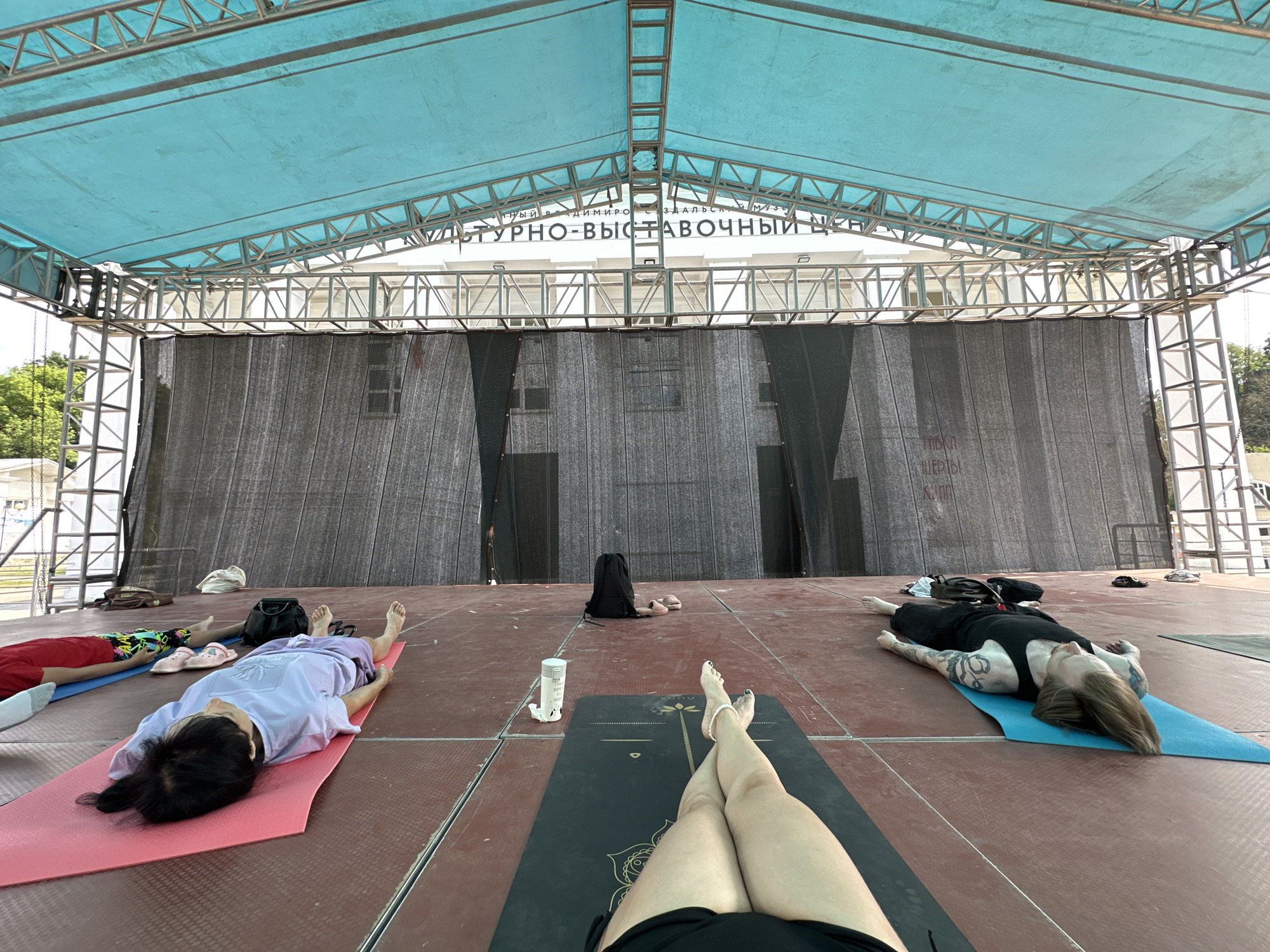 Four people are lying on yoga mats on a stage under a blue canopy. The stage floor is red. The people are in various poses, with their legs extended. One person is wearing a purple shirt and black pants, another is wearing a red shirt and patterned shorts, and a third is wearing a black tank top and black shorts. A fourth person's legs are visible in the foreground, wearing black shorts. There are bags and shoes scattered around the stage. In the background, there is a black curtain covering the stage and a sign with Cyrillic text. The scene appears to be outdoors. The sign in the background reads "КУЛЬТУРНО-ВЫСТАВОЧНЫЙ ЦЕНТР" which translates to "CULTURAL AND EXHIBITION CENTER", suggesting the location is likely the Cultural and Exhibition Center in Vladimir, Russia.