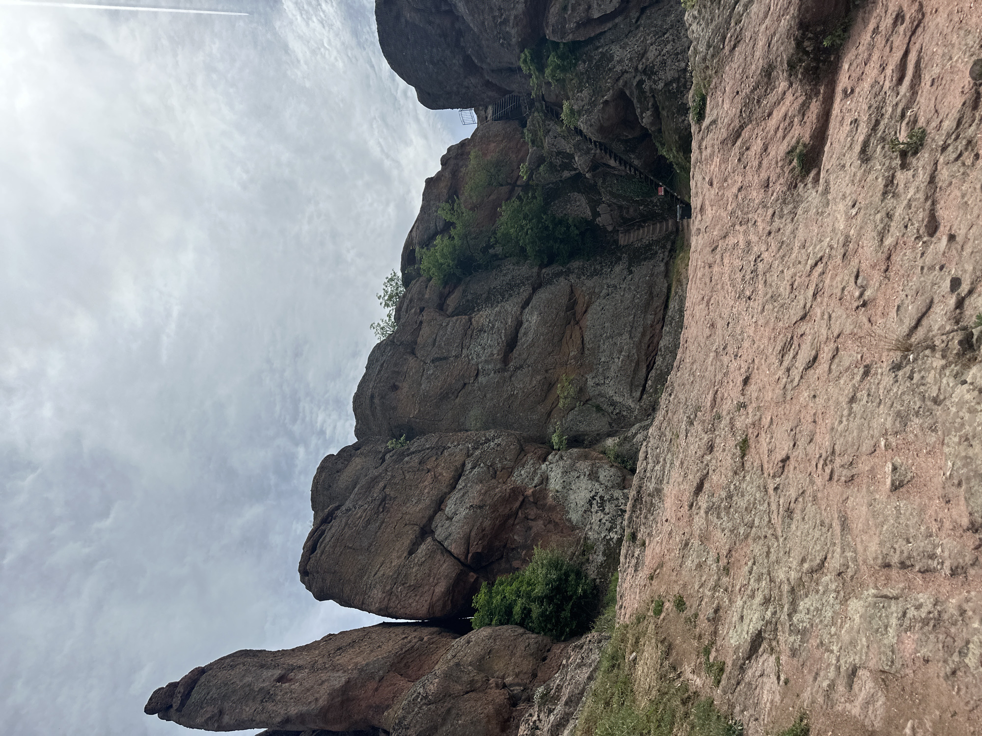 The image shows a low-angle shot of rock formations in Belogradchik, Bulgaria. The rocks are reddish-brown, weathered, and partially covered in vegetation. A set of metal stairs ascends a portion of the rock face, leading to a small platform. The sky is overcast with light, diffused clouds. A thin contrail is visible in the upper left of the frame.