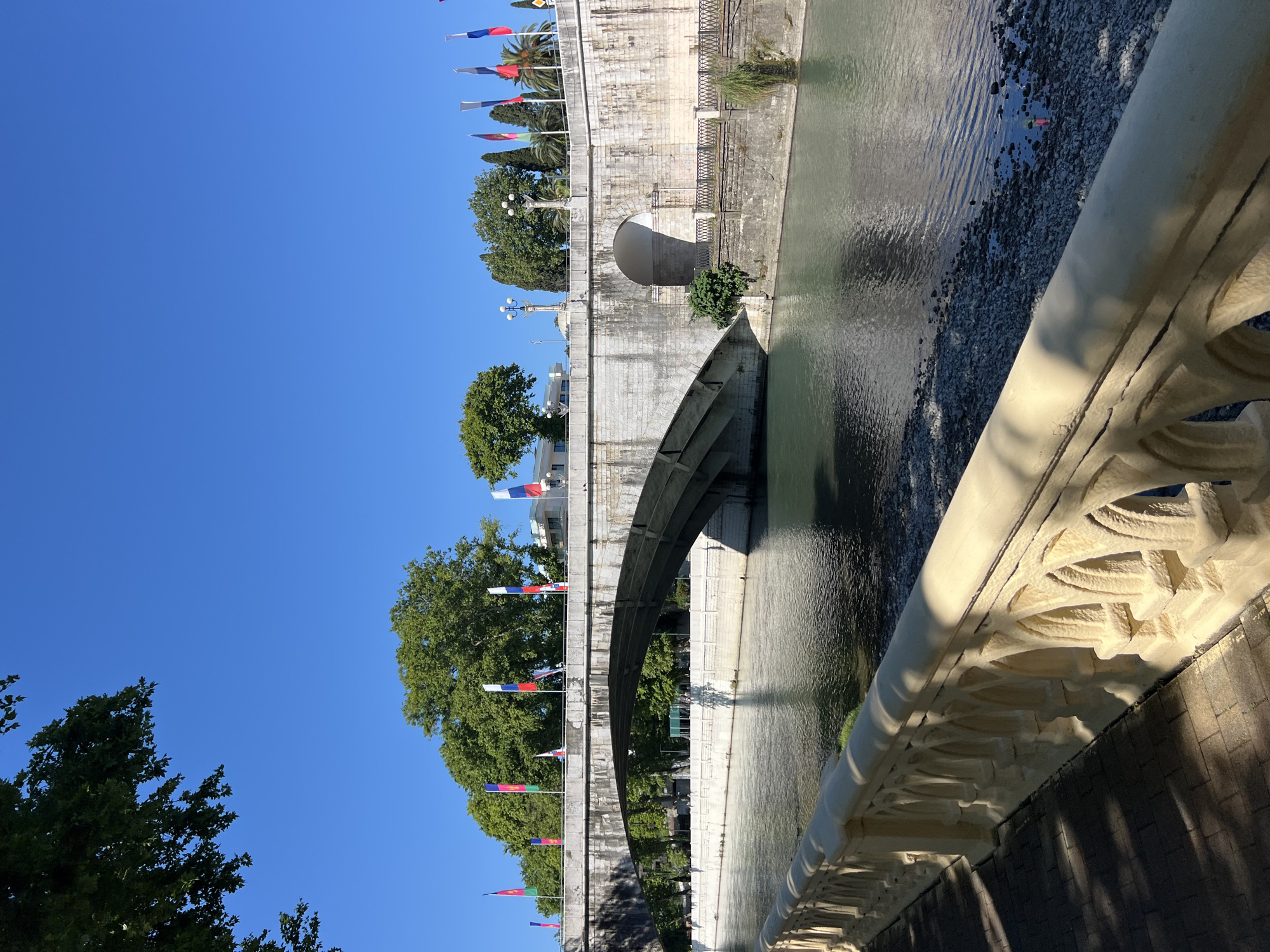 The image displays a view of a bridge over a body of water, likely a river, under a clear blue sky. The bridge is constructed of stone, with a large arch visible. Along the edge of the bridge, there is a decorative railing. Rows of flags, some appearing to be white, blue, and red, are strung along the side of the bridge, suggesting a celebratory or official occasion. Trees with lush green foliage are visible on the bank of the river and behind the bridge. The water is calm, reflecting the sky and the bridge. The sunlight casts shadows, indicating it is daytime, likely morning or afternoon. The scene is serene and appears to be outdoors in a city setting. There are no people clearly visible in the image, focusing instead on the architectural elements and natural surroundings. The flags might be a clue to the location or event, given the context of Sochi, Russia.
