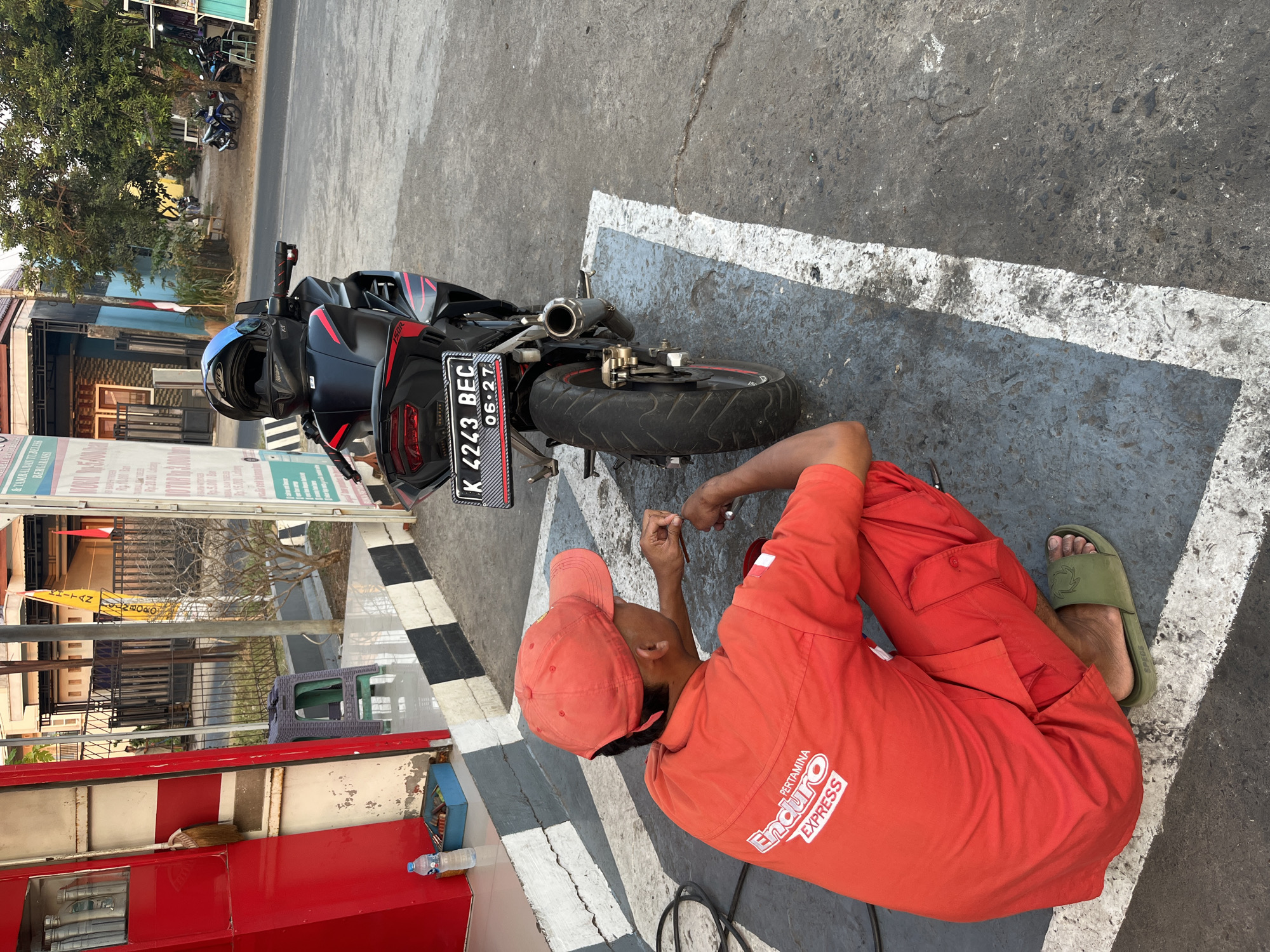 A male individual wearing an orange uniform, including a cap, and green slides, is crouched down next to a black motorcycle. The motorcycle has a license plate that reads "K 4243 BEC 06-27" and a blue and black helmet rests on its back. The individual appears to be attending to the motorcycle's rear tire. The scene is set on a concrete surface marked with white lines, suggesting a parking area or service bay. In the background, a red structure, possibly a kiosk or shop, is visible with various items displayed inside. The environment suggests an outdoor urban setting with buildings and trees in the distance, consistent with a location in Pecangaan, Indonesia. The lighting indicates it is daytime, with sunlight casting shadows.