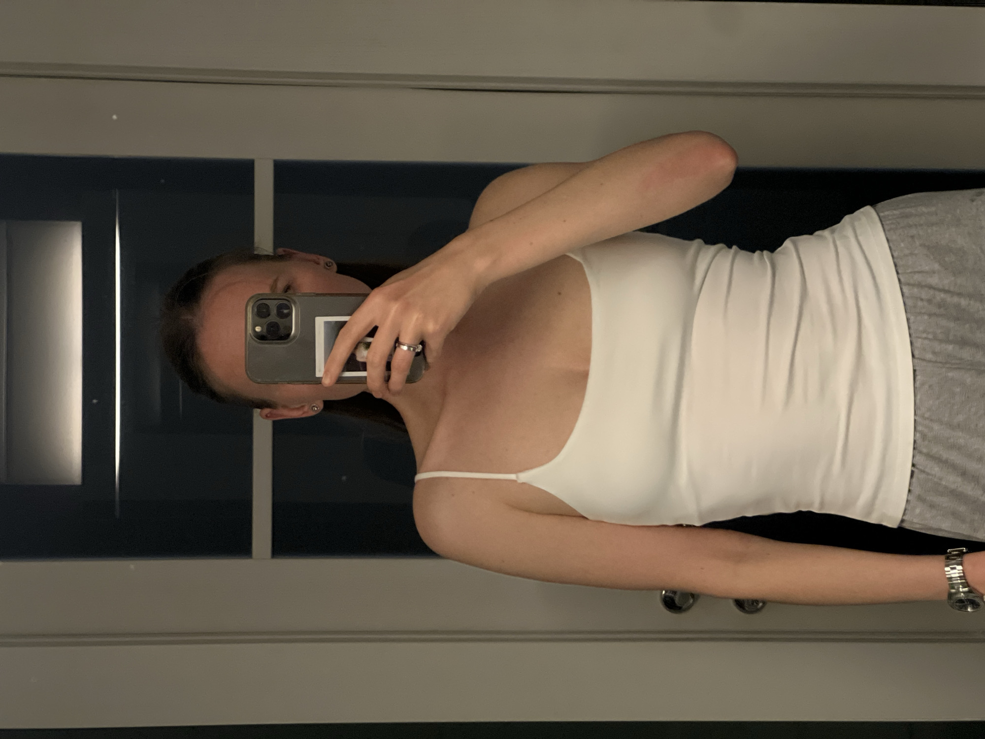 A woman is taking a selfie in a mirror. She is wearing a white tank top and gray sweatpants. She is holding an iPhone in her left hand and has a watch on her right wrist. The background is a gray and white paneled wall. The location is inside, likely a home or hotel room. There is no evidence to suggest the location is in Khimki, Russia.
