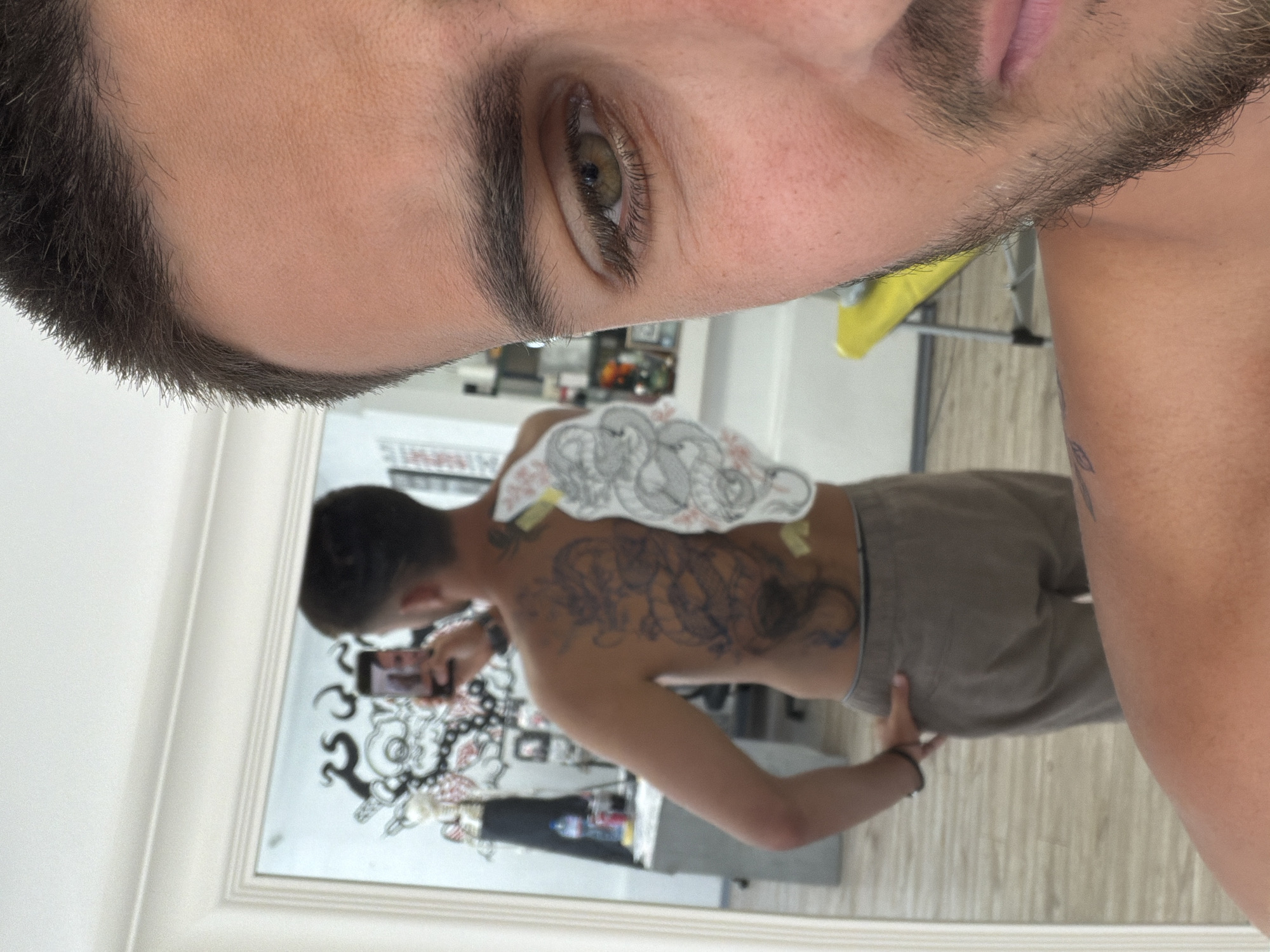 A man is posing for a mirror selfie in what appears to be a tattoo parlor. He is partially visible in the foreground, with his face cropped to show his right eye and part of his beard. In the background, reflected in a large mirror, the man's back is visible, showcasing a large, intricate dragon tattoo that is in progress. A stencil of the tattoo is also attached to his back. The tattoo parlor environment is visible in the reflection, with a desk, some bottles, and artwork on the walls. A yellow chair is also visible in the background. The man is wearing grey trousers and has a black bracelet on his left wrist. The image captures a moment of self-documentation during the tattoo process.