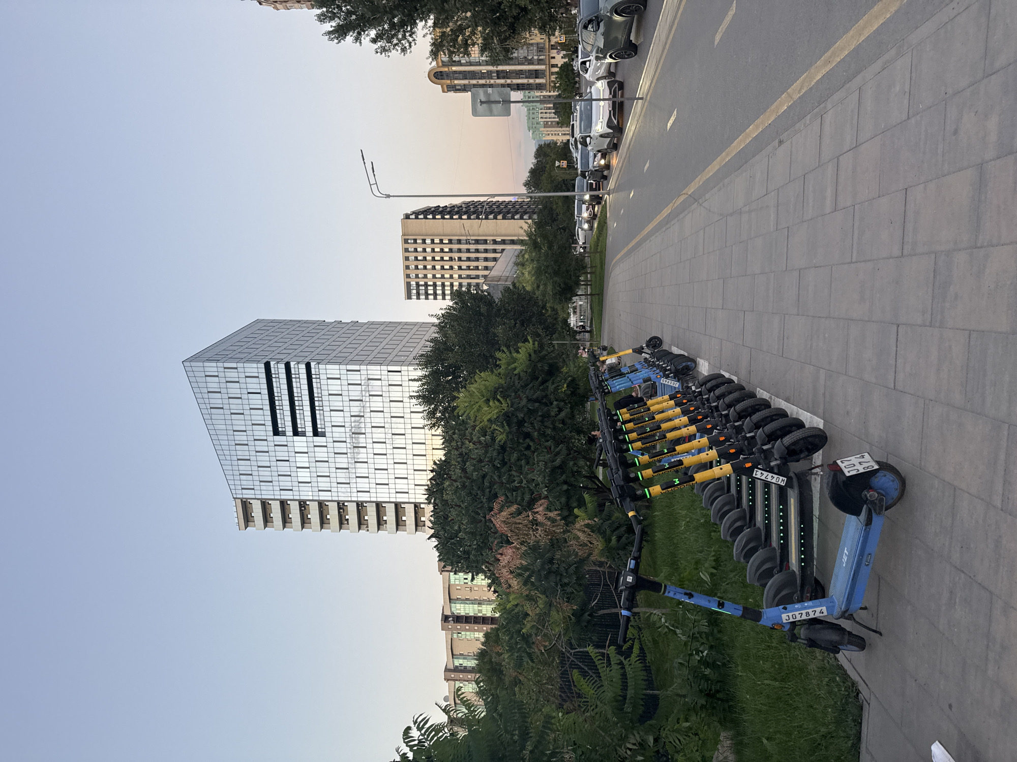 A street scene in Almaty, Kazakhstan, features a row of parked electric scooters on a paved sidewalk. The scooters, predominantly blue and yellow, are lined up neatly. Behind the scooters, lush green trees and shrubs form a natural barrier. In the background, modern high-rise buildings dominate the skyline, their glass facades reflecting the clear sky. A street with several cars is visible to the right of the sidewalk. The sky is a pale blue, suggesting it is either early morning or late evening.
