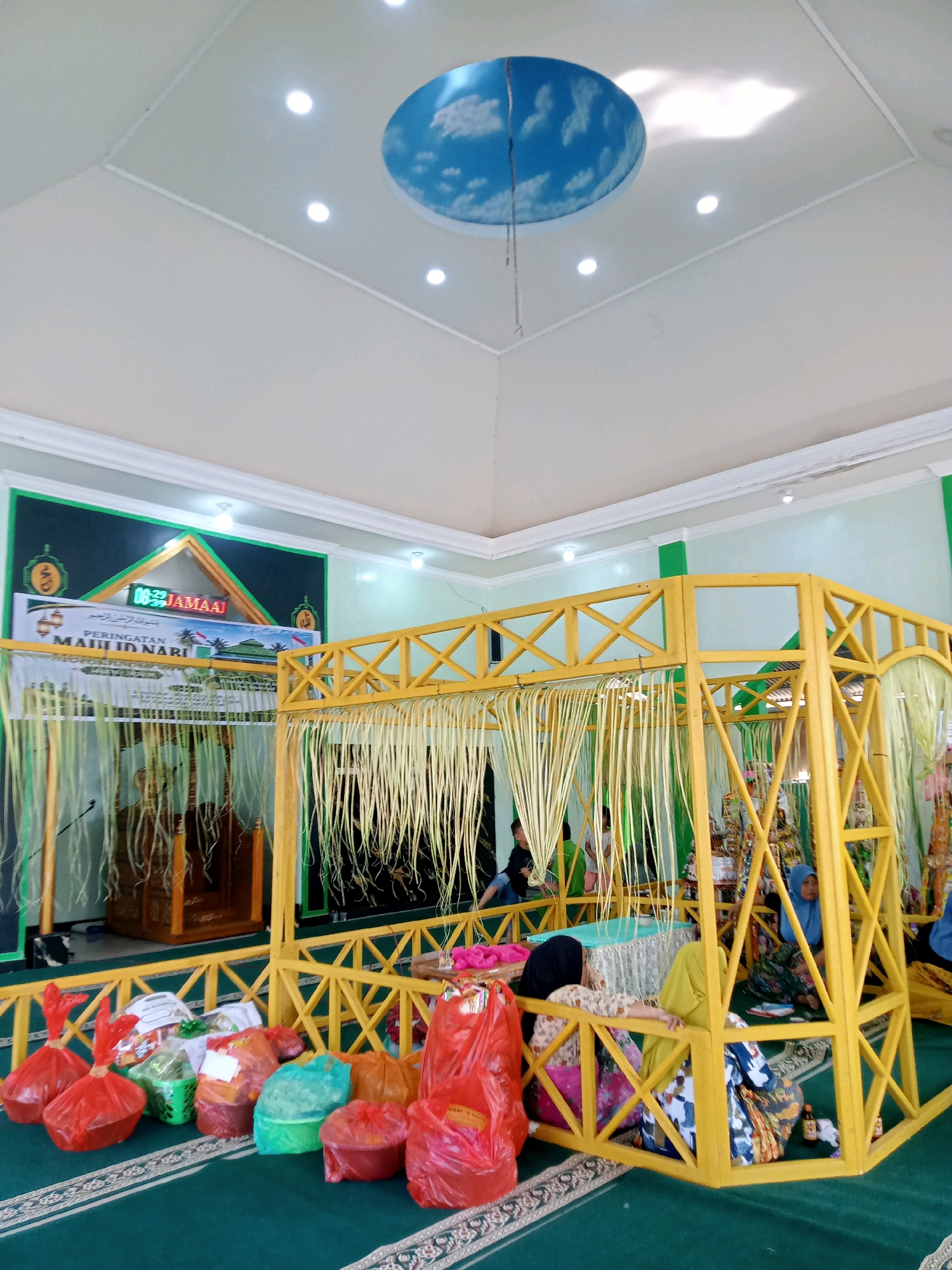 This image depicts an indoor event space in Mongolato, Indonesia, likely a mosque or community hall, decorated for a Maulid Nabi celebration. The room features a green carpet with a patterned border. In the foreground, several plastic bags filled with food items are arranged on the floor, some red, some green, and some orange. A prominent yellow wooden structure, adorned with hanging palm leaves, stands in the center of the room, serving as a decorative archway or stage. Behind this structure, a wooden pulpit is visible. The walls are painted in light green and white, with a large banner on the back wall featuring Arabic script and the text "PERINGATAN MAULID NABI." A digital clock displaying "00:22" is integrated into the banner. The ceiling is white with circular light fixtures and a circular decorative element painted with a sky and cloud motif. Several individuals, primarily women wearing hijabs and traditional clothing, are seated inside and around the yellow structure, suggesting they are participants or guests at the event. The overall atmosphere appears to be festive and communal.