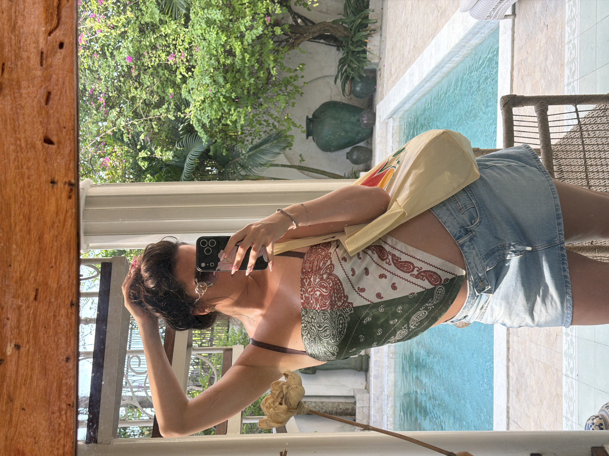 In North Kuta, Indonesia, a woman is captured in a mirror selfie taken outdoors. She is standing near a swimming pool with bright turquoise water, surrounded by lush green foliage and flowering trees. In the background, large, dark green ceramic vases and smaller pots are visible, adding to the tropical ambiance. The woman is wearing a bandana-style top with a paisley pattern in green, white, and red, and a light-wash denim mini-skirt. She has her hair pulled up and is wearing stylish sunglasses. She holds a smartphone in her right hand, taking the photo, and a light-colored tote bag hangs from her left shoulder. A wicker chair is positioned to her right, and the tiled patio and a decorative column are also in view. The scene is bright and sunny, reflecting the tropical setting.