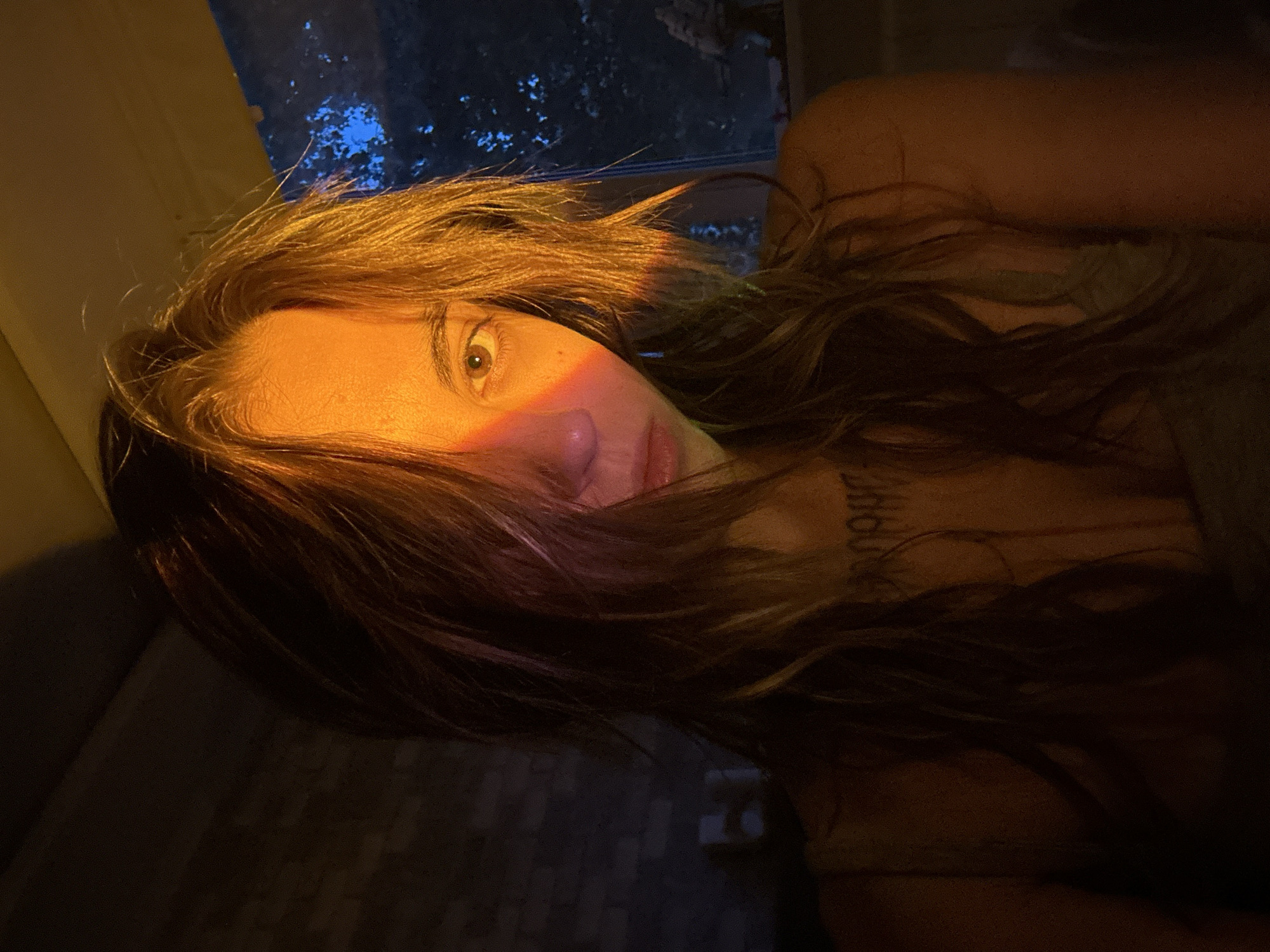 The image is a close-up, low-angle selfie of a person with long, wavy brown hair. The person's face is partially obscured by their hair, but their eye is visible, looking towards the camera. A rainbow-colored light stripe crosses their face, illuminating their cheekbone and eye. The background is dark and out of focus, with hints of trees and a blue sky visible through what appears to be a window. The lighting suggests it is either dawn or dusk. The person is wearing a dark-colored top, and a tattoo is visible on their collarbone. The overall mood is intimate and atmospheric. Based on the provided location context, the scene is set in Moscow, Russia.