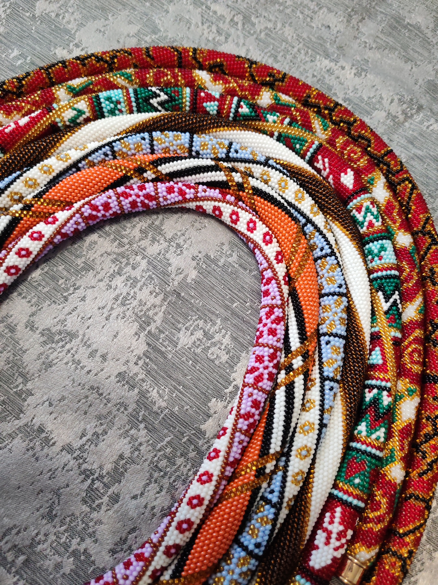 A close-up, top-down shot showcases a collection of intricately beaded necklaces arranged in a semi-circular fashion on a textured gray surface. The necklaces display a variety of vibrant patterns and colors, including red, white, orange, purple, blue, green, gold, and black beads. Some necklaces feature geometric designs, while others have floral motifs. The lighting highlights the glossy texture of the beads and the detailed craftsmanship of each piece. The background surface appears to be a fabric or a textured material with a distressed, abstract pattern. There are no people or signs of a specific city or location visible in the image.