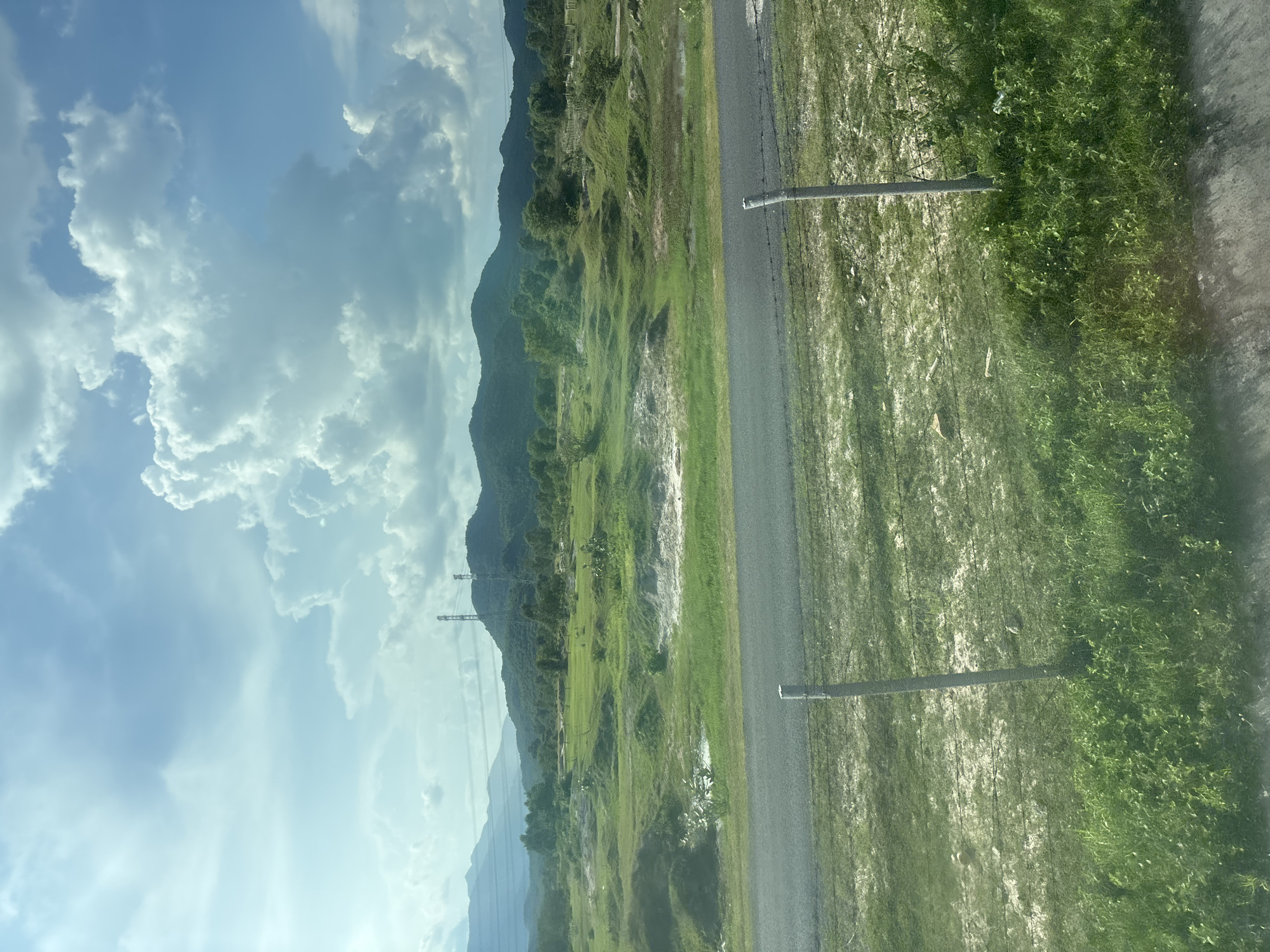 The image depicts a rural landscape under a partly cloudy sky. In the foreground, a paved road runs horizontally across the frame, bordered by grassy verges. Two grey posts with barbed wire are visible in the middle ground, suggesting a fence line. Beyond the road, a vast expanse of uneven green terrain is dotted with small mounds and patches of lighter soil. In the distance, rolling, tree-covered hills form the horizon. Two tall, lattice-style electricity pylons stand prominently on the side of the hills. The sky is a mixture of bright blue and fluffy white clouds, with a hint of sunlight breaking through. The overall impression is of a serene, natural setting with man-made infrastructure integrated into the landscape.