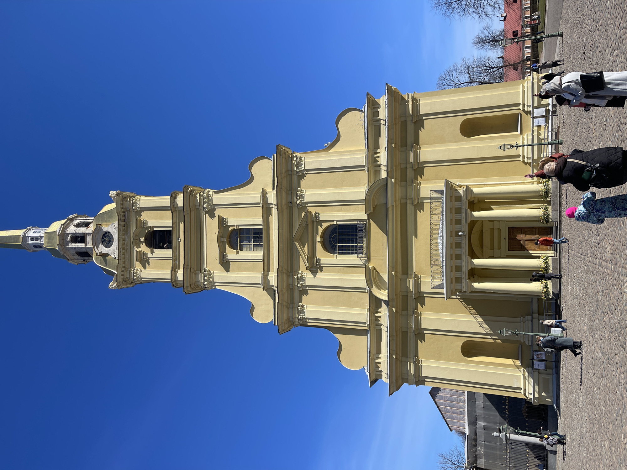 A full shot, slightly rotated, features the Peter and Paul Cathedral in Saint Petersburg, Russia under a clear blue sky. The cathedral is yellow with ornate detailing and a tall spire. Several people are standing in the foreground, near the cathedral's entrance. The ground is cobblestone. Bare trees are visible in the background.