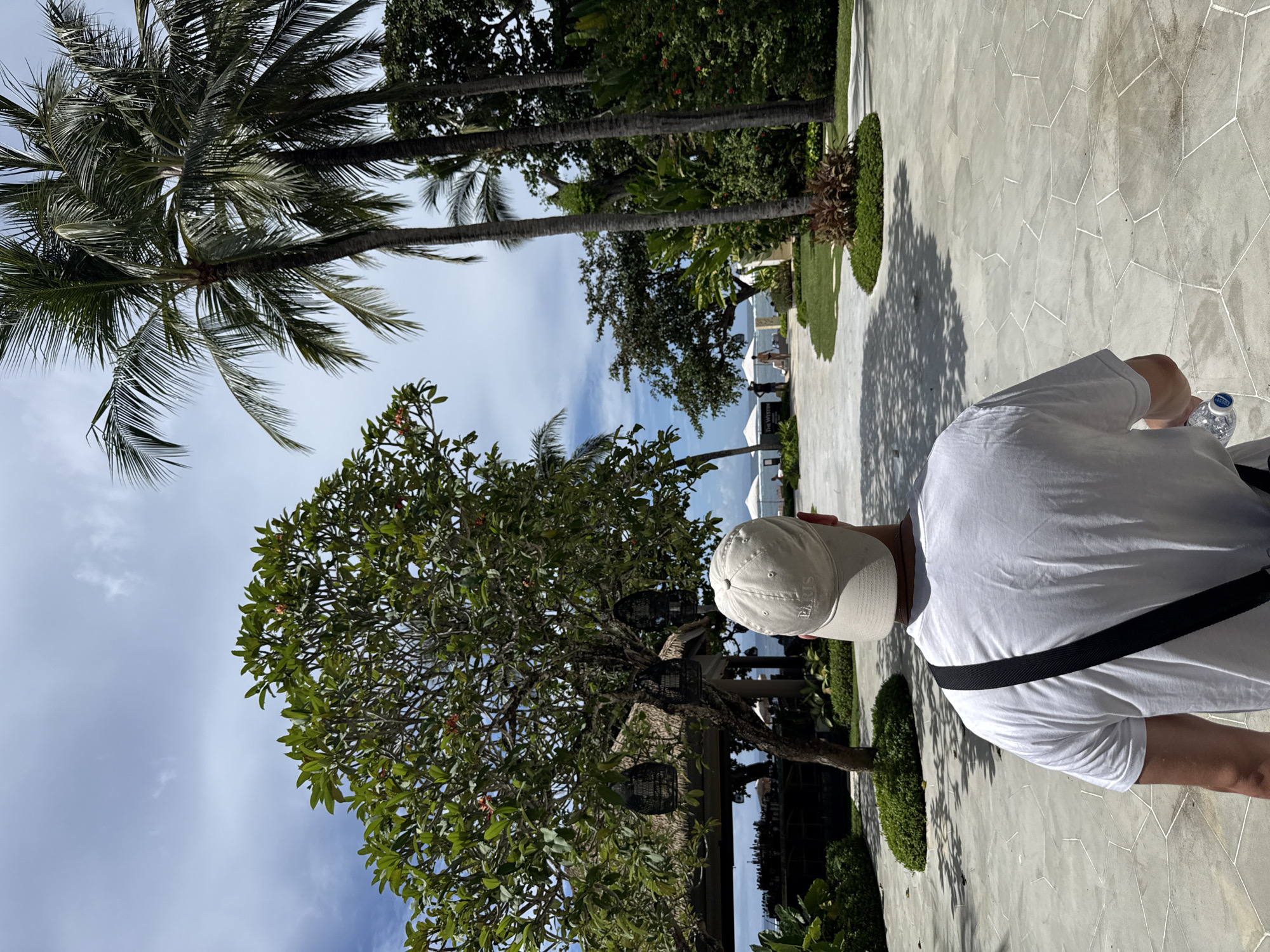 A man wearing a white t-shirt and a beige cap stands on a paved walkway in Kuta Selatan, Indonesia. The walkway is made of irregular, polygonal stones. Palm trees and other tropical vegetation are visible. Further in the background are a building and a glimpse of the ocean. The sky is partially cloudy.