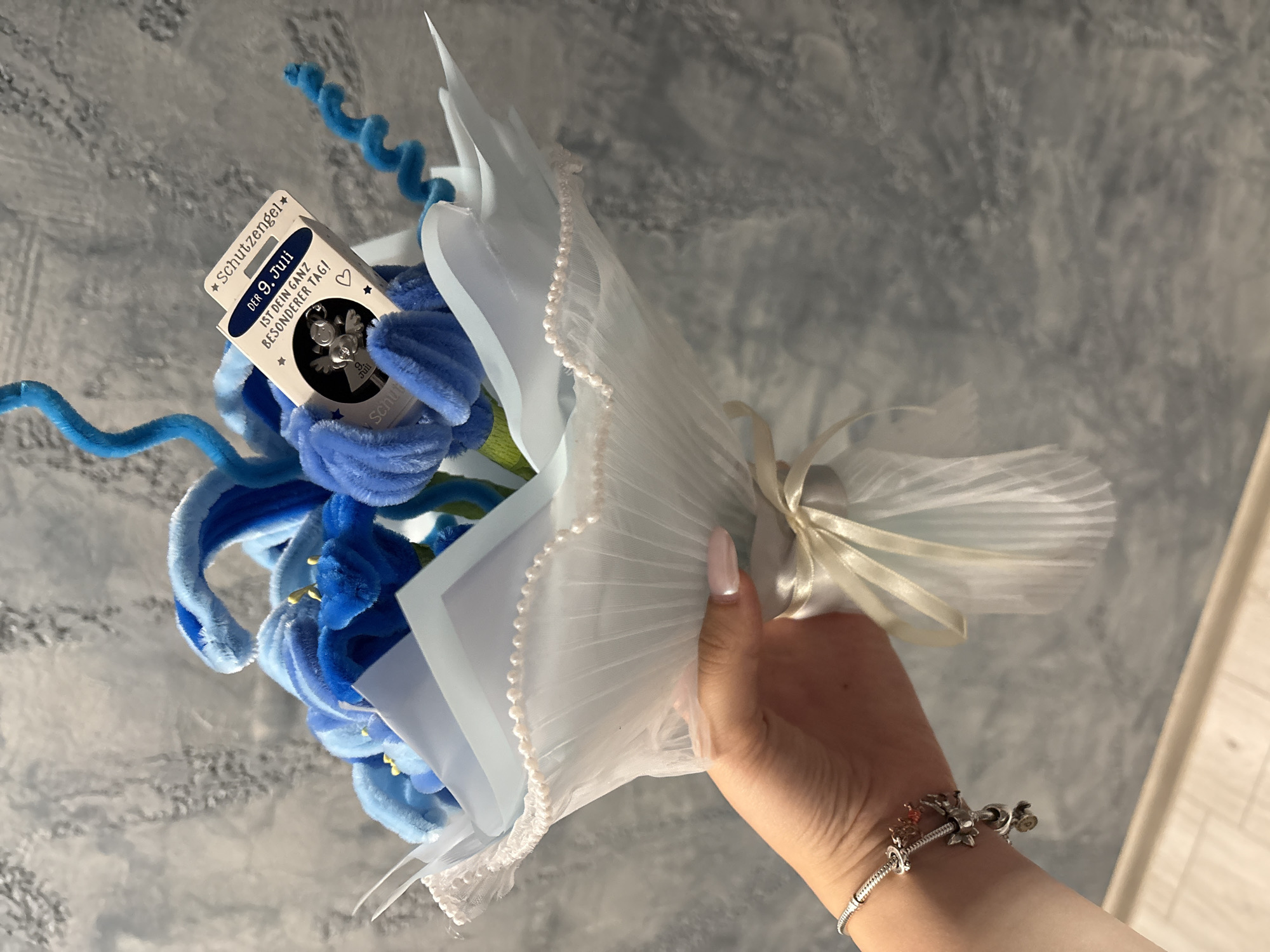A hand is holding a bouquet of blue pipe cleaner flowers wrapped in light blue tulle and tied with a cream ribbon. A small cardboard tag with a silver angel charm is attached to the bouquet. The tag reads "Schutzengel" and "DER 9. JULI IST DEIN GANZ BESONDERER TAG!". A silver bracelet with charms is on the wrist of the hand. The background is a gray, textured wall.