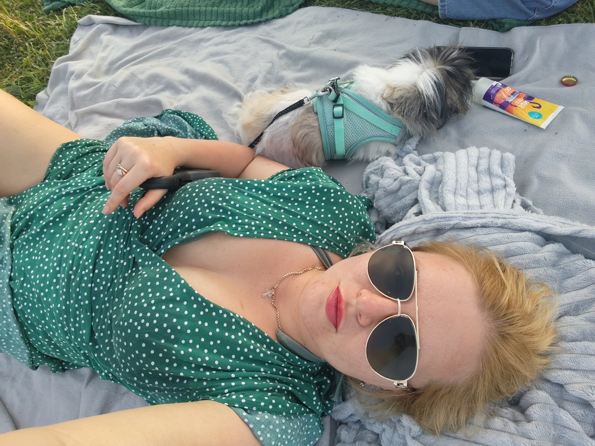 In Podshivalovo, Russia, a woman with blonde hair and sunglasses lies on a grey blanket in a grassy area, wearing a green polka-dot dress. She is holding a dark object, possibly a phone, in her right hand. A small dog wearing a teal harness lies next to her. A tube of sunscreen and a bottle cap are also on the blanket.