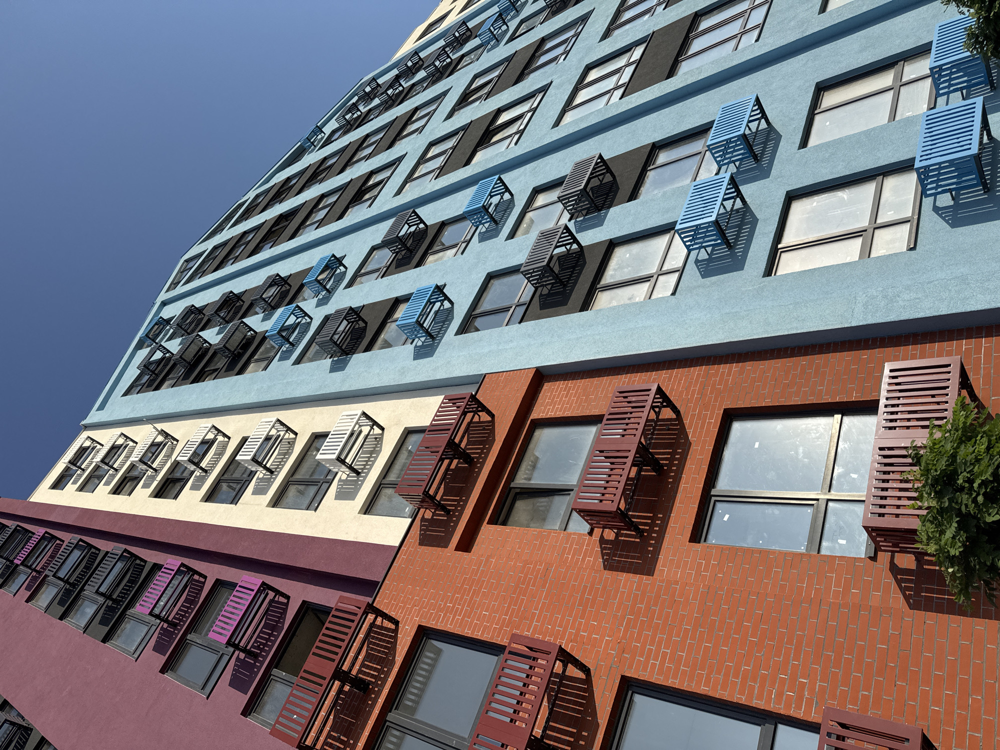 The image shows the exterior of a modern, multi-story apartment building in Yevpatoriya, Ukraine. The building features distinct sections painted in different colors: a deep purple on the left, followed by a light yellow, and a prominent light blue on the right. The architecture is characterized by a repetitive pattern of windows and external air conditioning units mounted on metal brackets. These brackets are colored red on the purple and yellow sections, and black or blue on the blue section. Some windows are open, revealing the interior, while others are closed. The building is seen from a low-angle perspective, looking upwards towards a clear blue sky, suggesting a sunny day. Shadows cast by the air conditioning units and balconies are visible on the building's facade. In the lower right corner, some greenery is present. The scene appears to be a residential area with a focus on the building's facade and its architectural details.