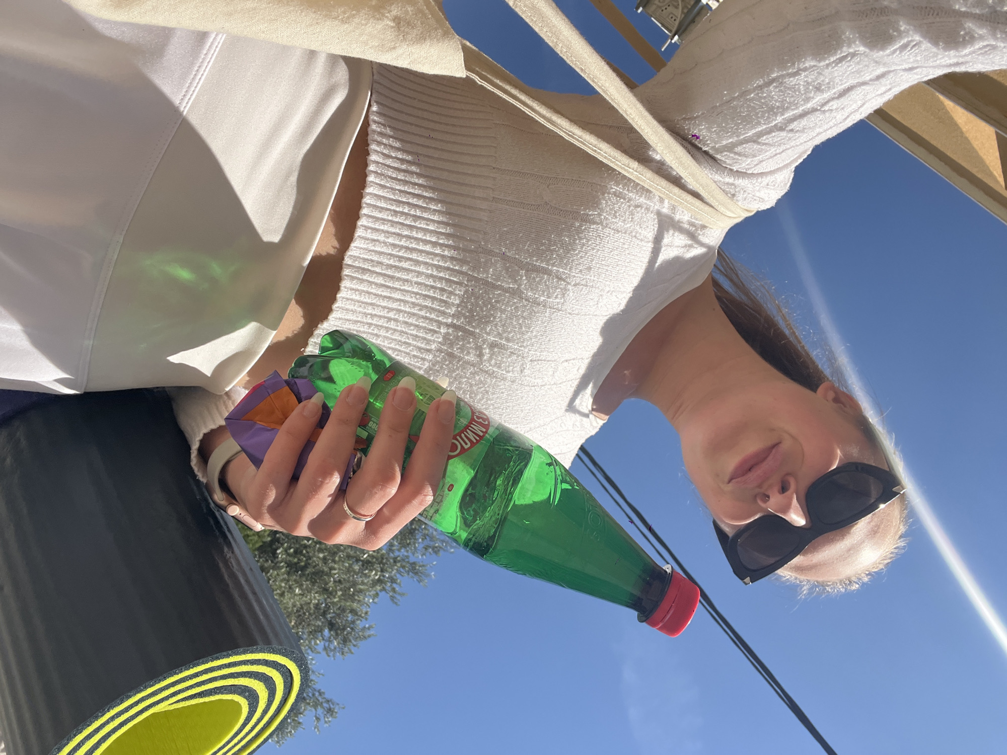 The image shows a low-angle outdoor selfie taken in Boreti, Montenegro, under a clear blue sky. A person, likely female, is captured from the chest up, wearing sunglasses and a white textured, long-sleeved top. They hold a green plastic bottle with a red cap in their right hand, along with crumpled colorful wrappers (purple and orange). A silver ring is visible on their finger, and a light-colored watch or wristband is on their left wrist.
In the lower left foreground, a black yoga mat with distinctive bright yellow layered edges is rolled up. The sky is intensely blue and clear, indicating bright daylight. A strong sun flare is visible on the right side of the frame, and shadows are cast on the person's white clothing, with a green reflection from the bottle also visible on the fabric. In the background, an olive tree can be seen, along with faint architectural details of a building. The person's face is partially obscured by sunglasses, but a relaxed expression is suggested.
On the label of the green bottle, the brand name "GUSINJKA" is clearly visible, along with numerical markings such as "200" and "000". The overall scene suggests an outdoor activity or exercise session, possibly yoga, on a sunny day.