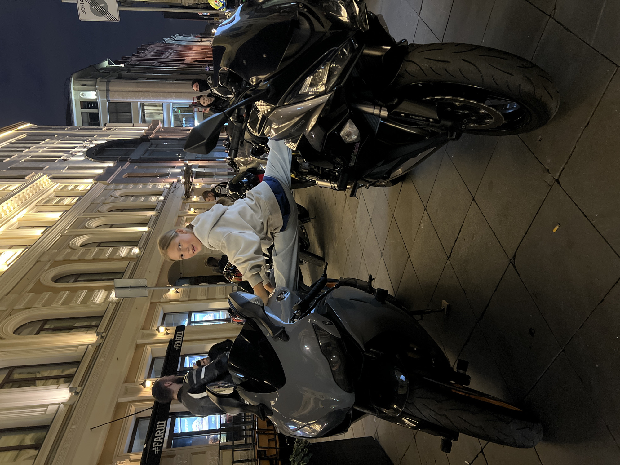 A young blonde girl is sitting on a black motorcycle, looking at the camera. She is wearing a light gray hoodie and light blue pants. Her right hand is on the handlebars. Another motorcycle, also black, is parked behind her. A third motorcycle, light gray, is parked in front of her. The scene is a street at night, with buildings lining the sidewalk. The buildings appear to be old and ornate, with arched windows and decorative trim. Some of the windows are lit from within. There are streetlights visible, casting a warm glow on the scene. The ground is paved with square tiles. Visible text includes "#FARII" on banners in the background. The overall mood is casual and relaxed, with the girl appearing playful and engaged with the camera.