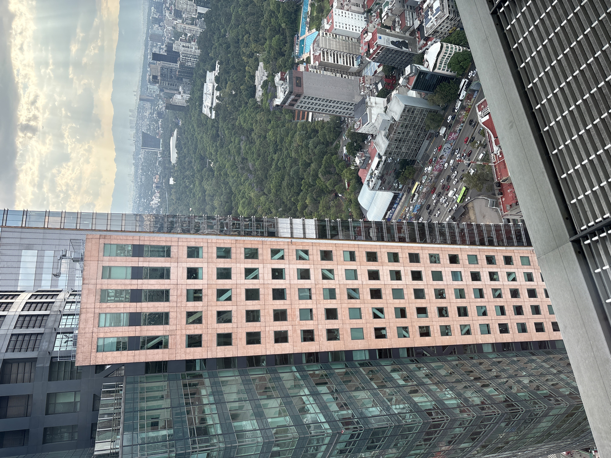 This high-angle shot, taken from what appears to be a skyscraper in Mexico City, Mexico, features a built environment with a mix of architectural styles. Below the point of view are the roofs of adjacent buildings, including a pinkish building with multiple small square windows, a modern glass structure, and a gray building with a metal grid-like façade. In the distance, a vast cityscape extends to the horizon, partially obscured by haze. A large green space, likely Chapultepec Park, is visible amidst the buildings. The street traffic is visible along a road bordering the park. The sky overhead is overcast with scattered clouds.