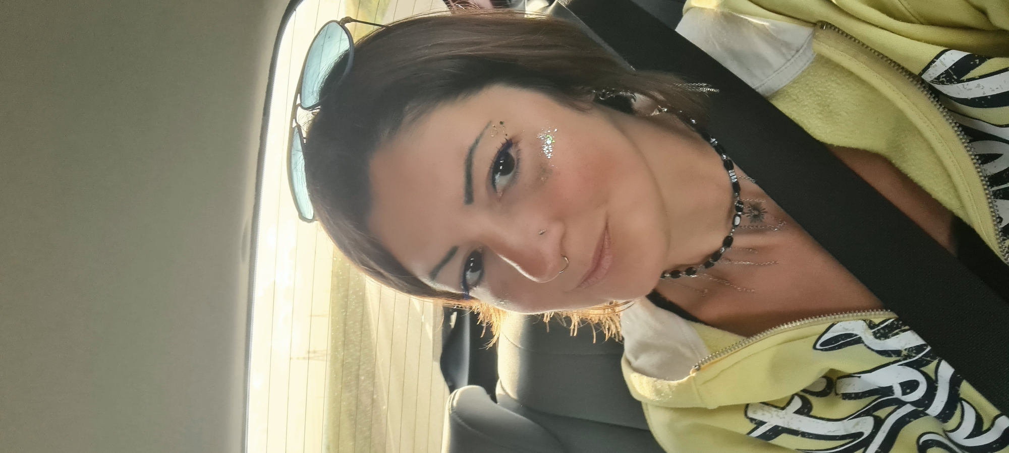 A woman with glittery makeup and a nose ring is photographed indoors, likely in a car given the visible seatbelt and window. She is wearing a yellow zip-up hoodie and a beaded necklace. Sunglasses rest on top of her head. The background shows blurred daylight and glimpses of the exterior, possibly a road. The location appears to be within Bnei Shimon Regional Council, Israel, as the image was sourced from there.