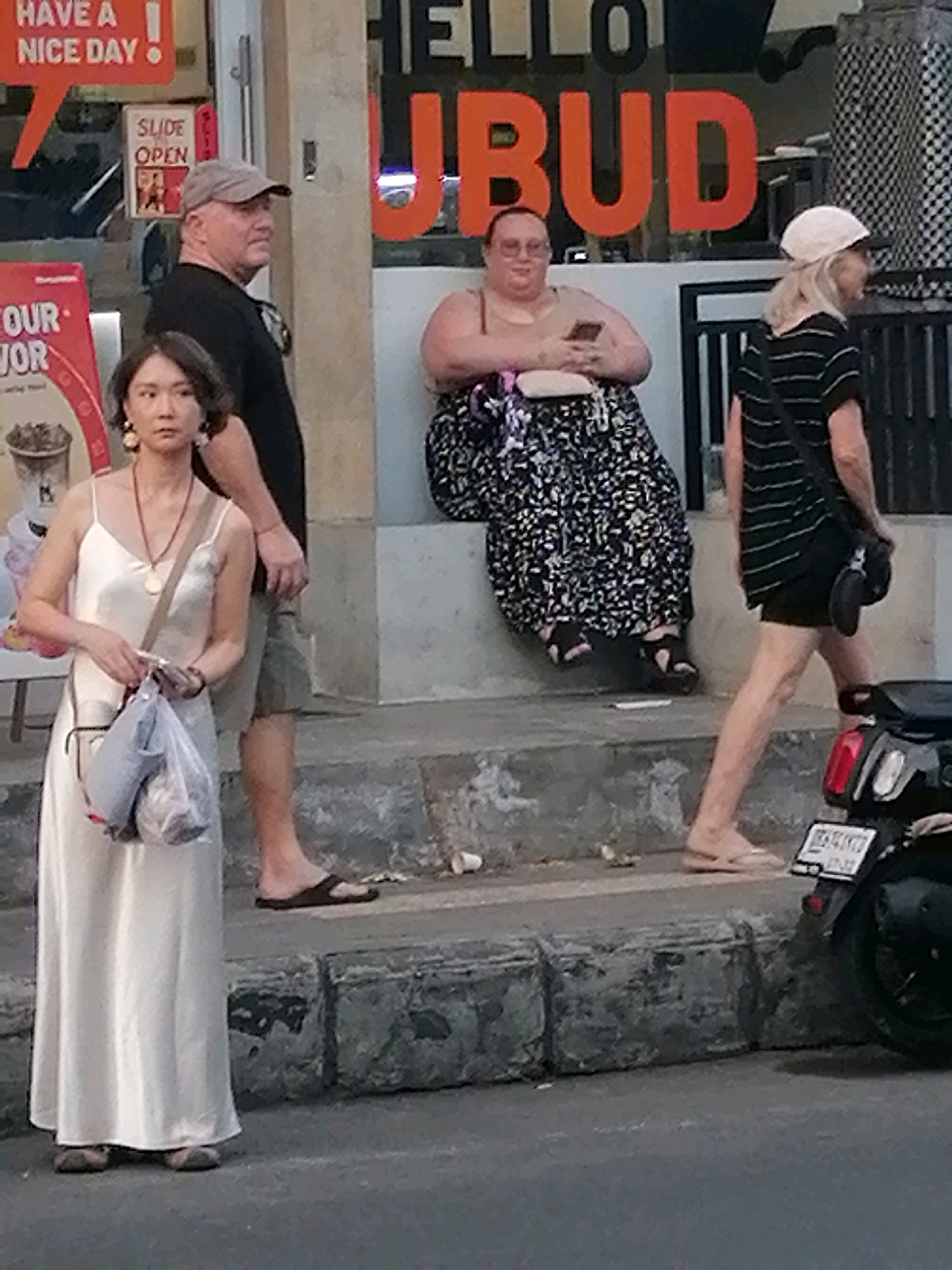 The image shows a street scene in Ubud, Indonesia, with several people visible. In the foreground, a woman wearing a long, cream-colored dress and a necklace stands on the street, holding a plastic bag. To her right, a man in a grey baseball cap and black t-shirt is partially visible, looking to his right. In the background, a person wearing glasses and a beige top sits on a ledge, holding a mobile phone and wearing a patterned skirt. To the far right, another person in a black and white striped t-shirt and a white hat walks away from the camera, carrying a black bag. The storefront behind the people features large orange lettering that spells out "UBUD". A scooter is parked on the right side of the frame. The ground appears to be paved, with some concrete steps and debris visible.