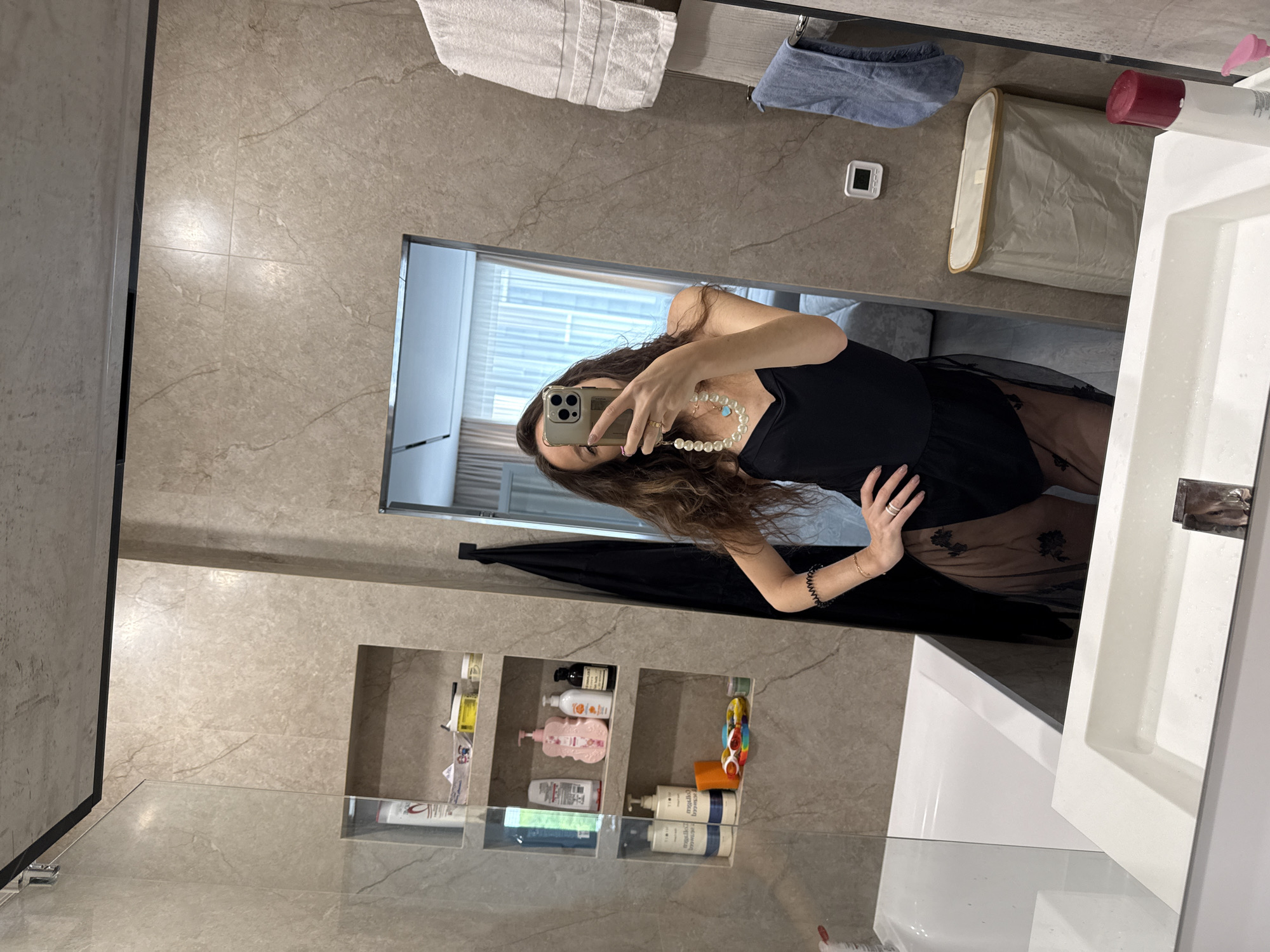A mirror selfie taken in a bathroom in Khimki, Russia, shows a person with long brown hair, wearing a black top and sheer bottoms with a floral pattern, holding a smartphone. The bathroom has beige marble-patterned walls, built-in shelves with toiletries, a white sink, and a shower with a glass door. A window is visible in the background, possibly with curtains. A hamper, towels, and a digital thermometer are on the counter.