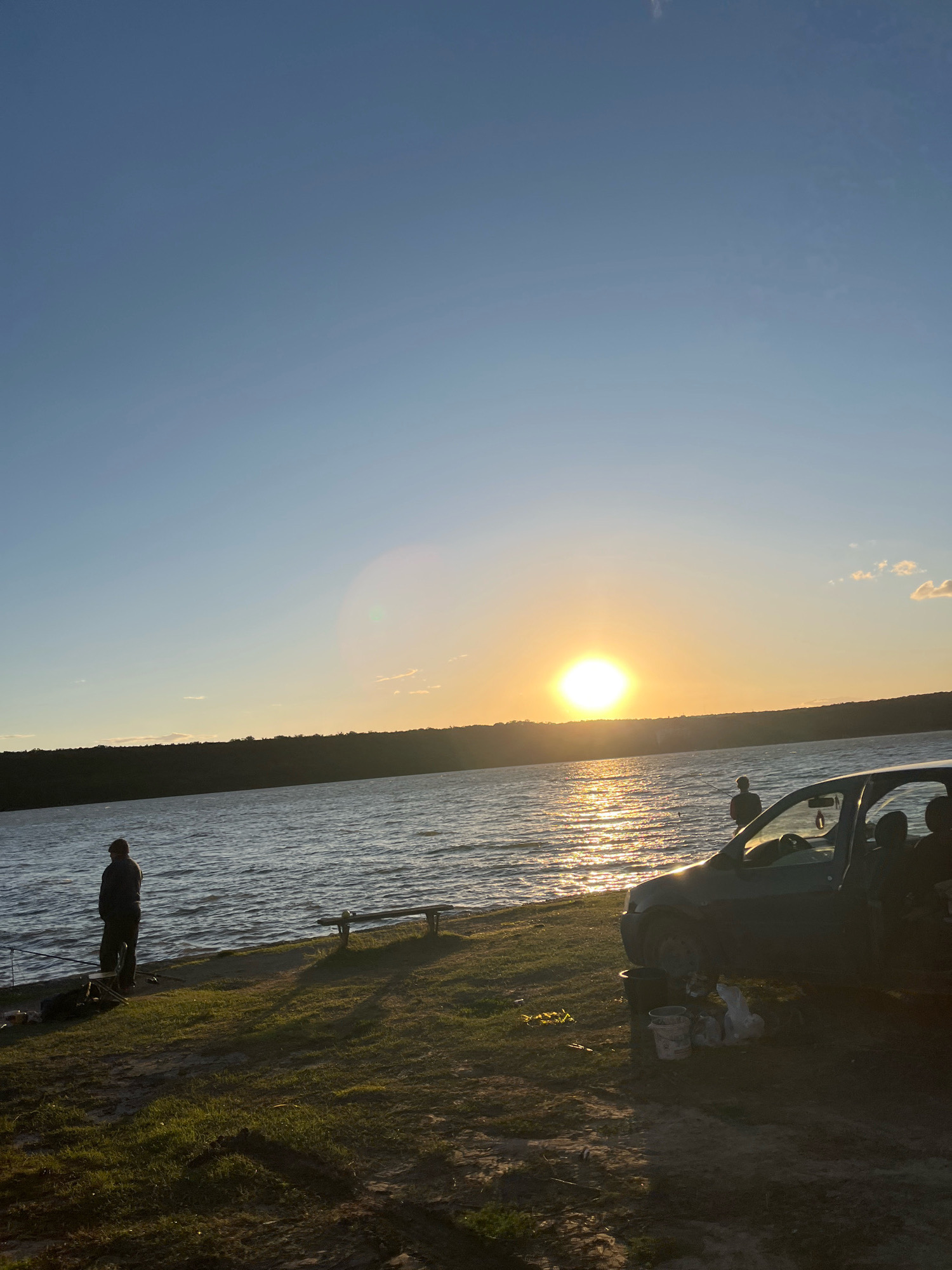 The image depicts a serene sunset over the Dniester River near the village of Cocieri, Moldova. The sky is a gradient of clear blue at the top transitioning to a warm orange and yellow hue near the horizon, where the sun is setting. The sun's reflection creates a shimmering path across the water.
On the grassy bank of the river, a person stands fishing, silhouetted against the water. Further down the bank, a parked car with its door open is visible, with at least two individuals inside. Scattered around the car are buckets and bags, suggesting a fishing or recreational outing. A wooden bench is also present on the bank, offering a place to rest. The overall environment is one of tranquil natural beauty with human activity present.