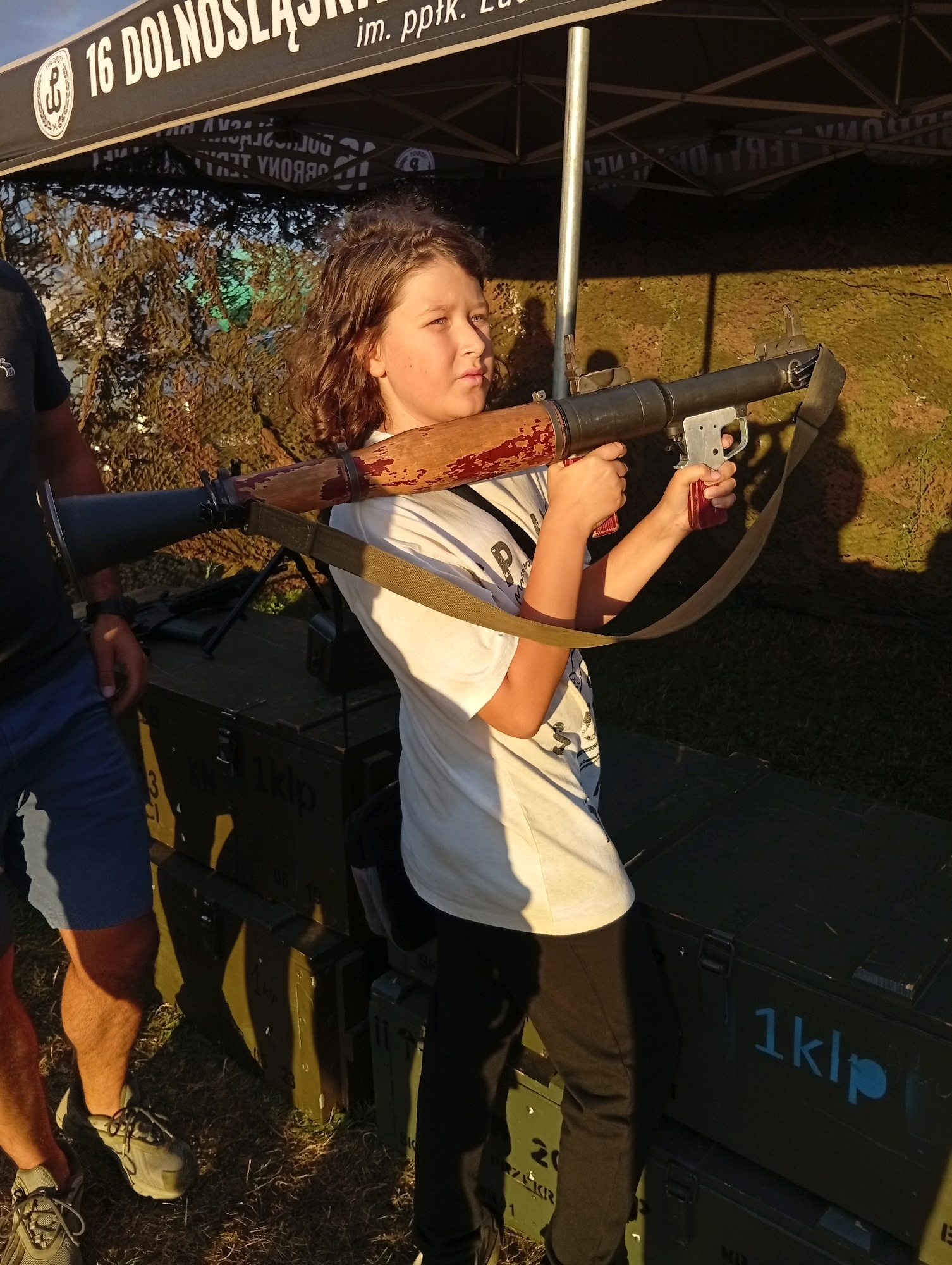A young boy with curly brown hair, wearing a white t-shirt and black pants, stands outdoors holding a replica rocket-propelled grenade launcher. He is looking to his right, with his left hand gripping the weapon's pistol grip and his right hand near the trigger. The launcher is resting on his right shoulder, with a green strap slung across his chest. Behind him, several dark green ammunition crates are stacked. The crates have markings, with "1klp" visible on one of them. A man's legs, wearing blue shorts and grey athletic shoes, are visible on the left side of the image. The scene appears to be set up in an outdoor area, possibly at an event or fair, with a tent in the background that has a banner with Polish text, including "16 DOLNOŚLĄSKIE". The lighting suggests it is late afternoon or early evening, with long shadows cast on the ground. The location is identified as Szewce, Poland.
