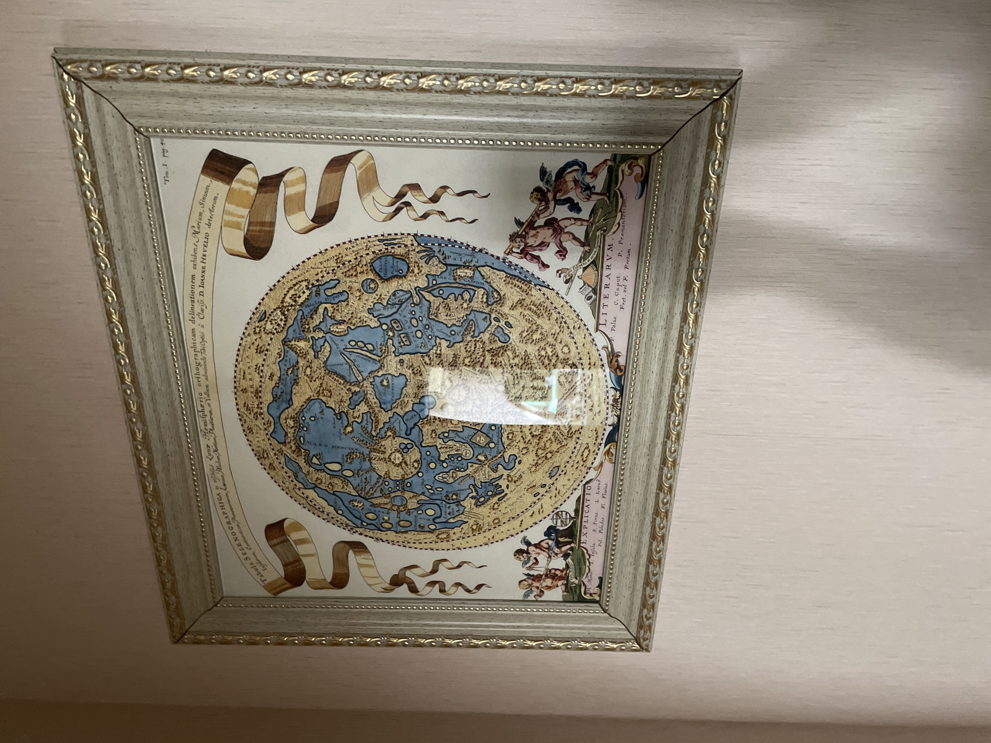 A framed print hangs on a wall, depicting a vintage map of the moon's surface. The map is circular and shows the lunar landscape in blues and browns, with decorative scrolls at the top and bottom. Tiny figures are depicted on the sides, seemingly supporting the map. The print's frame is light gray with gold detailing. Text in Latin surrounds the map. The wall is a light, neutral color, possibly light beige or off-white. There is no indication in the image that the location is Moskovskiy, Russia.
