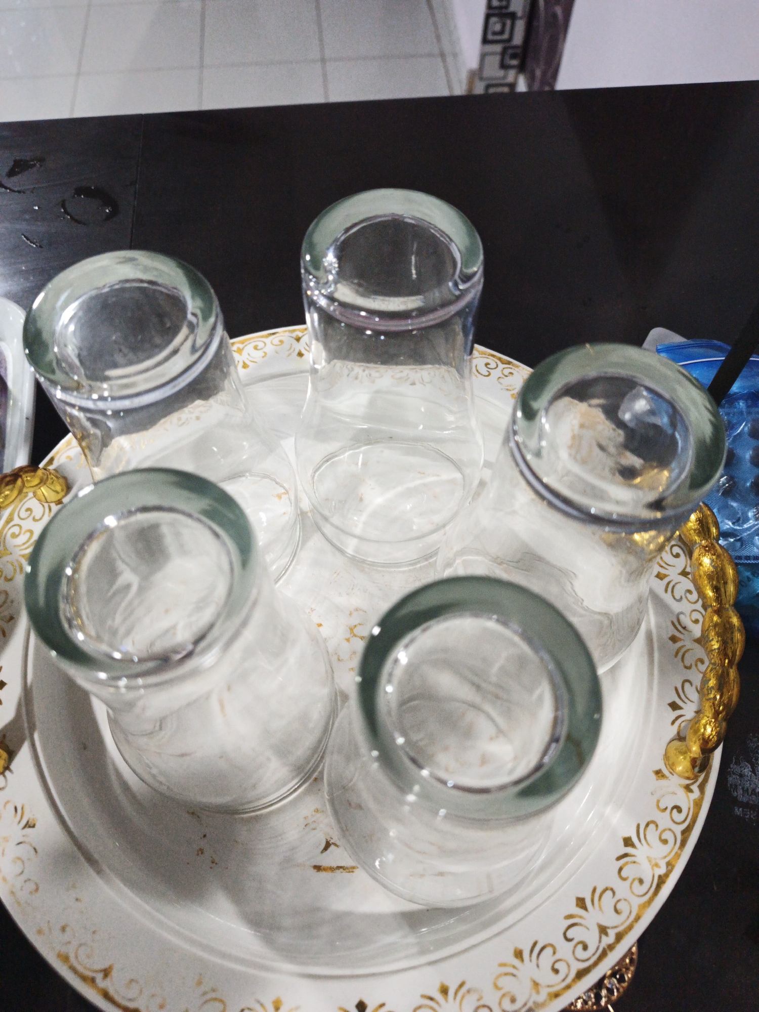 The image depicts a close-up, overhead view of five empty clear glass tumblers placed upside down on a white tray. The tray has ornate gold detailing along its rim and edges, featuring a floral or scroll pattern. The glasses are arranged in a circular fashion on the tray. The tray itself sits on a dark, flat surface, possibly a table, with a tiled floor visible in the background. To the right of the tray, a blue plastic container is partially visible, along with a dark object, possibly a straw. The lighting suggests an indoor setting. There are no people or animals visible in the image, and no specific actions or interactions are taking place. The scene is static. The location is identified as Kelapapati, Indonesia.