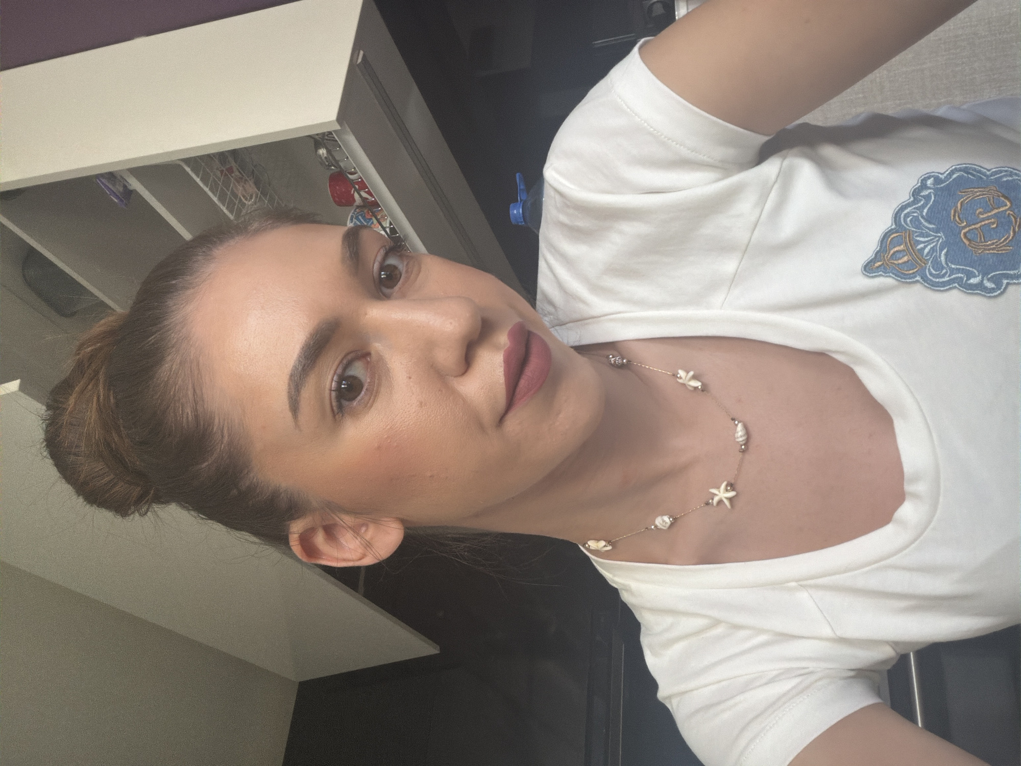 The image shows a close-up, selfie-style shot of a woman with brown hair pulled up in a bun. She is wearing a white t-shirt with a necklace that features shell-like pendants and beads. She has makeup on, including lipstick. The background appears to be an indoor setting, possibly a kitchen or living area, with white cabinets and shelves visible. The lighting suggests it's daytime. The image does not provide any specific information or visual cues that would allow for its location to be identified within the city of Sofia, Bulgaria.
