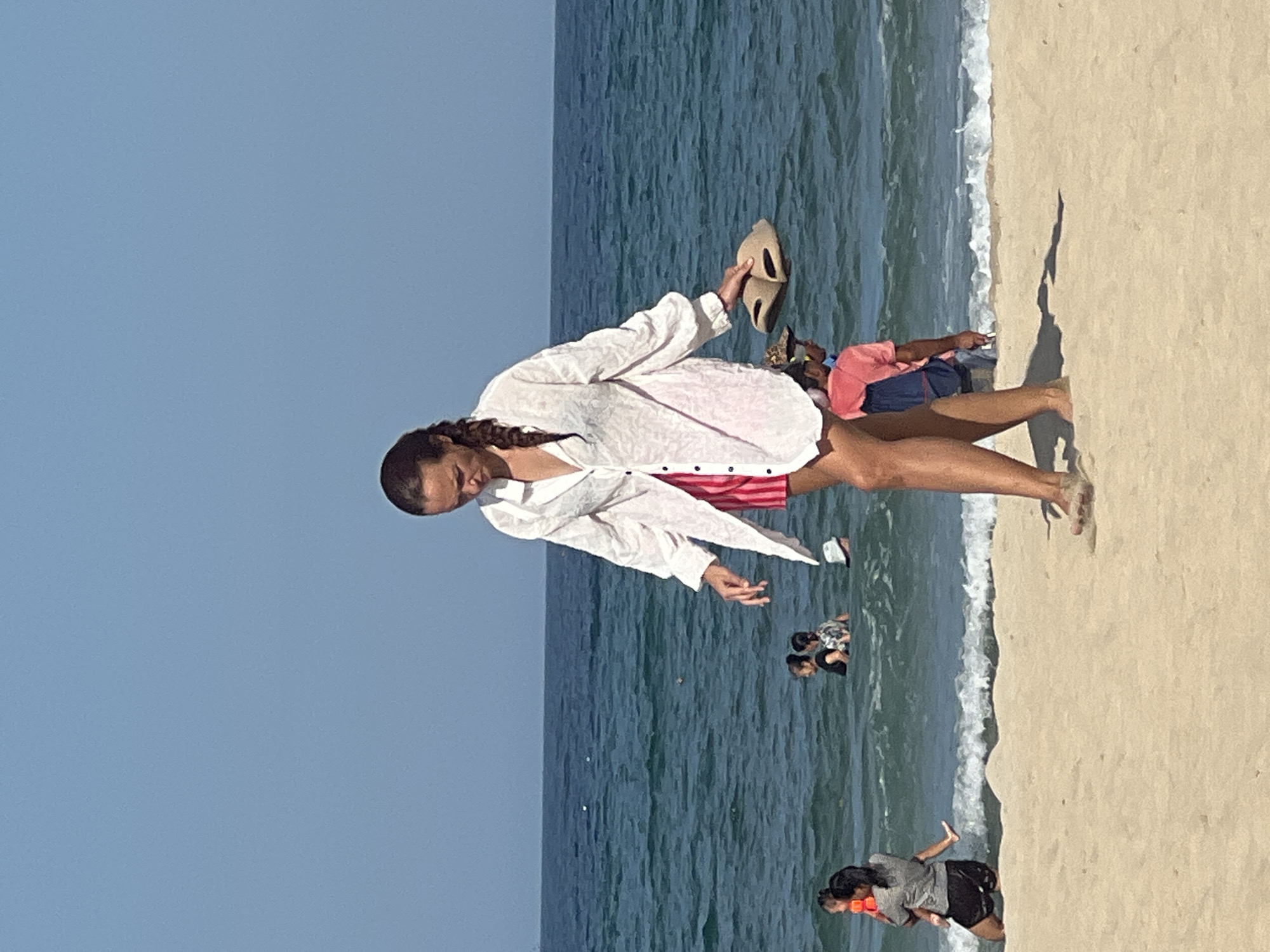 At a beach in Da Nang, Vietnam, a woman with braided hair walks across the sand towards the camera, carrying a pair of tan sandals in her right hand. She is wearing a loose-fitting, long-sleeved white shirt over striped pink and white shorts. Her head is bowed as she looks down at her feet, which are dusted with sand. Behind her, the blue ocean stretches to the horizon under a clear sky, with waves breaking gently on the shore. Several people are visible in the water, some wading and others swimming. To the left, a woman in a grey shirt and black shorts holds a baby wearing an orange life vest. Further back in the water, two women are visible, and to the right of the woman in the foreground, a person in a pink shirt with a blue backpack walks away from the camera. The sand in the foreground is light beige, showing footprints and shadows cast by the woman and another person.