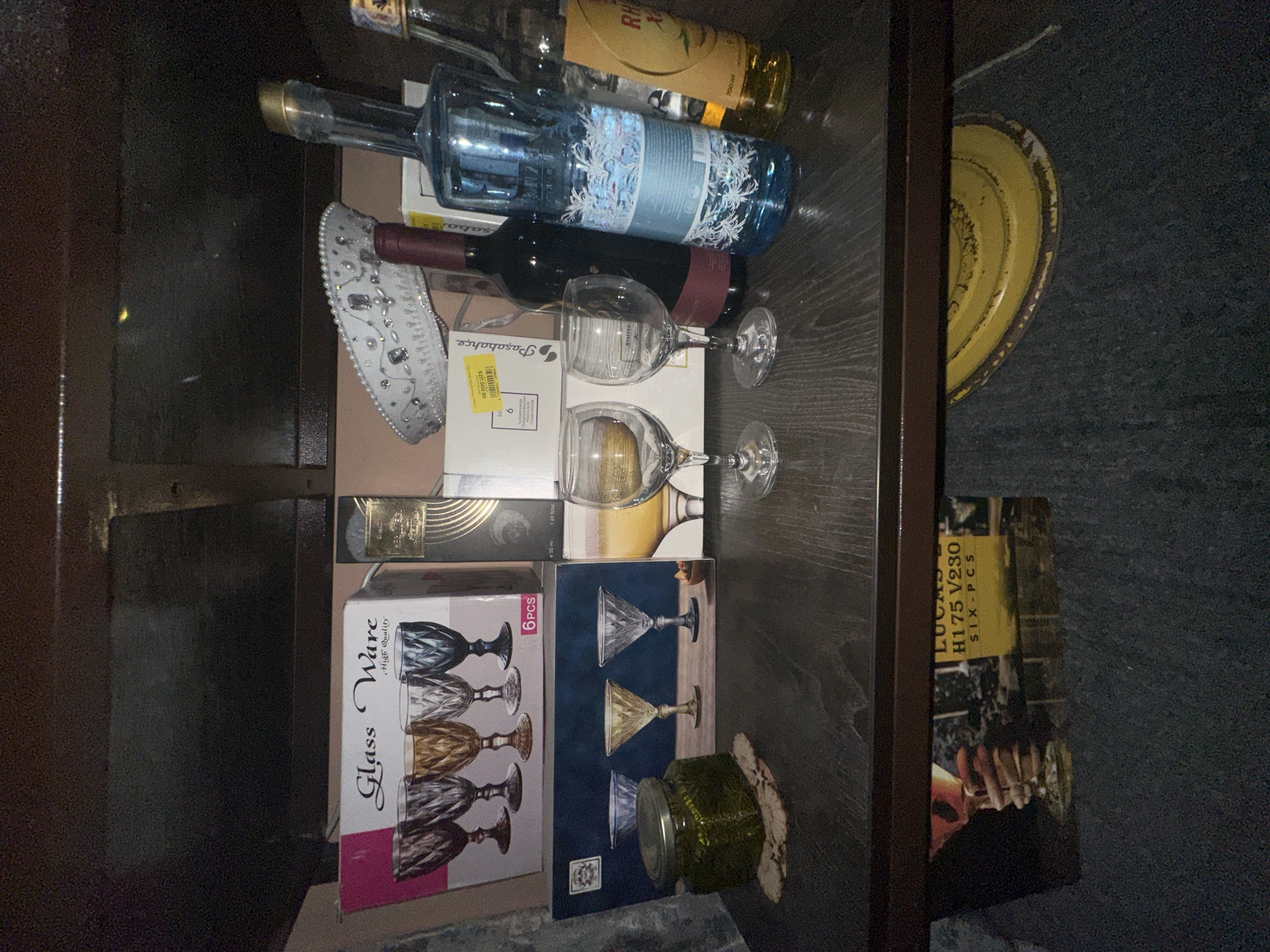 This media file captures a close-up, overhead view of a collection of items stored in a dark wooden drawer or cabinet. The scene is dimly lit, suggesting an indoor setting possibly at night or in a poorly lit room.
The primary subjects are various bottles, glassware, and some packaging. Among the items are several bottles of alcohol, one with a blue label featuring an ornate white design, and another with a yellow label. Two wine glasses are positioned on top of some packaging. Also visible is a decorative white item with beadwork and embellishments, possibly a hat or a decorative piece.
Further down in the drawer, there are boxes containing glassware. One box is labeled "Glass Ware High Quality" and shows an illustration of faceted wine glasses. Another box, with a blue background, also depicts martini-style glasses. Next to these boxes is a small green glass jar. A yellow circular object, possibly a plate or lid, is partially visible on the right side of the frame.
The visible text includes brand names and product information. Text on one box reads "Paşabahçe" and "46 000 UZS". Another label states "LUCAS H175 V230 SIX-PCS". There is also text indicating "700 ml" and "e 50 ml 1.69 fl/oz.". The overall impression is a storage space filled with assorted decorative and beverage-related items.